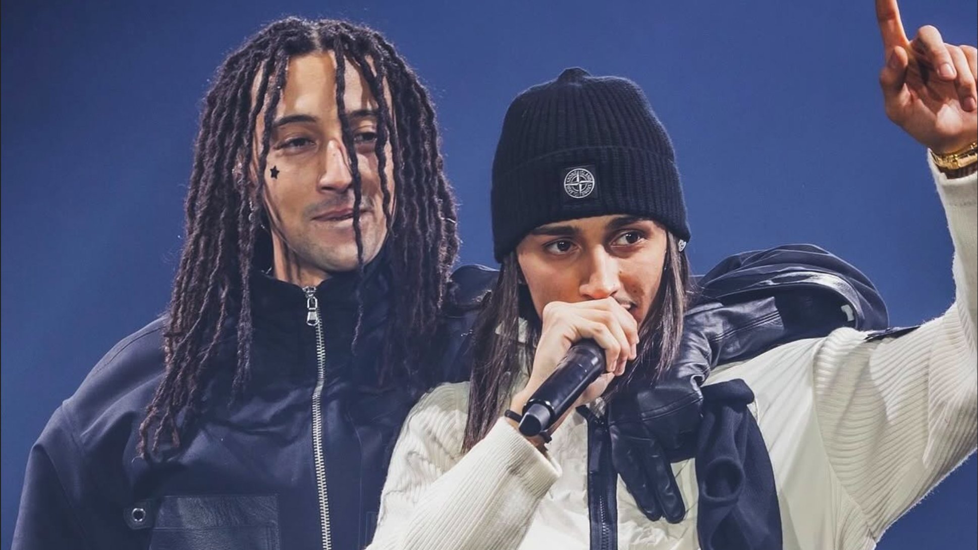 Due artisti sul palco: uno con capelli ricci e l'altro con un cappello nero, entrambi in abbigliamento sportivo.