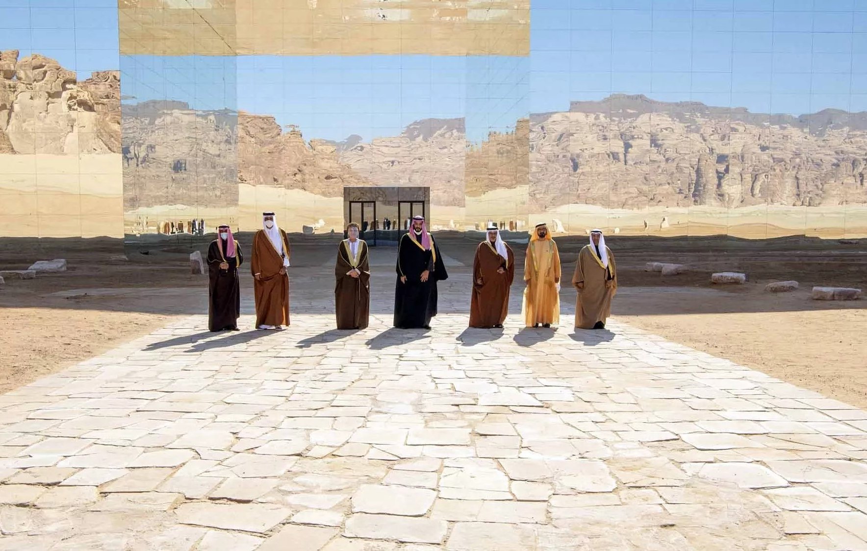 L'immagine mostra un gruppo di uomini vestiti con abiti tradizionali, dimostrando un'atmosfera di formalità e celebrazione. Sono disposti in fila su una pavimentazione di pietre chiare, con un paesaggio desertico sullo sfondo. Dietro di loro si erge una struttura riflettente che sembra integrarsi con l'ambiente circondante, creando un gioco di luci e riflessi. Le montagne in lontananza aggiungono profondità alla scena, conferendo un senso di grandiosità e atmosfera unica.