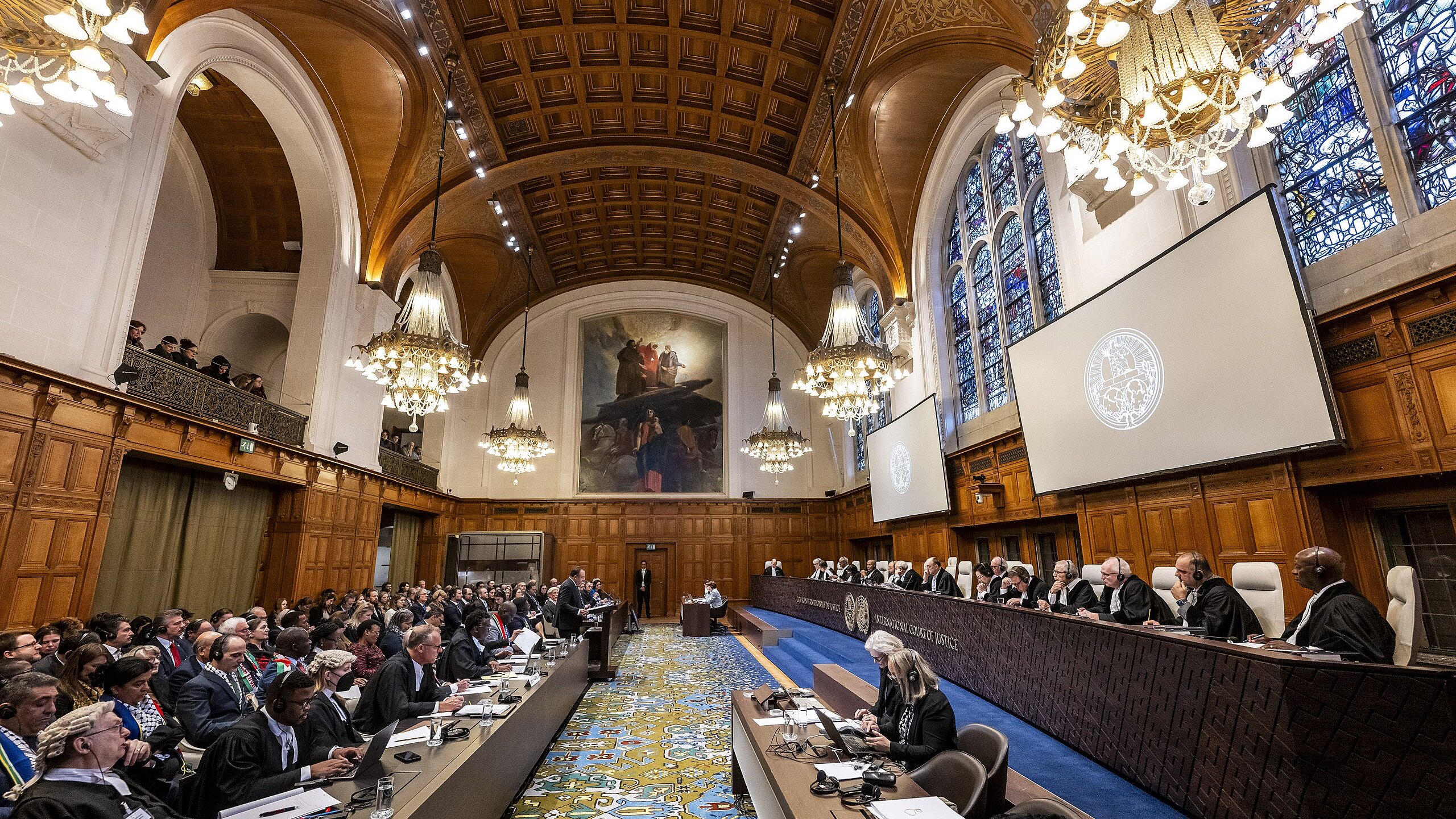 L'immagine mostra un'aula di giustizia, probabilmente una corte internazionale. La sala è caratterizzata da un'architettura imponente con soffitti alti, lampadari eleganti e finestre ad arco che lasciano filtrare la luce. In primo piano, c'è un lungo tavolo con diversi avvocati e rappresentanti, mentre sul lato opposto, una serie di giudici sono seduti su una piattaforma rialzata. Il pubblico è composto da molte persone, alcune delle quali prendono appunti. L'atmosfera sembra formale e rispettosa, tipica di un'importante udienza legale.