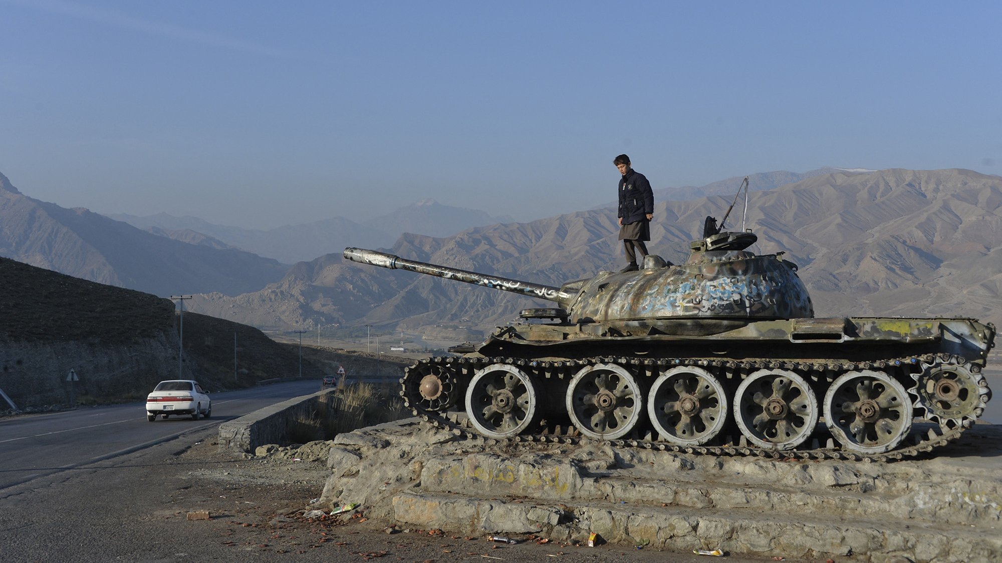 L'immagine mostra un carro armato, probabilmente un T-55, posizionato su una base con un giovane che si trova sopra di esso. Sullo sfondo si possono vedere le montagne, che creano un paesaggio arido e suggestivo. La strada accanto al carro armato ospita un'auto che viaggia, mentre il cielo è sereno e blu. L'atmosfera è di calma, ma il carro armato evoca un passato di conflitto e guerra.