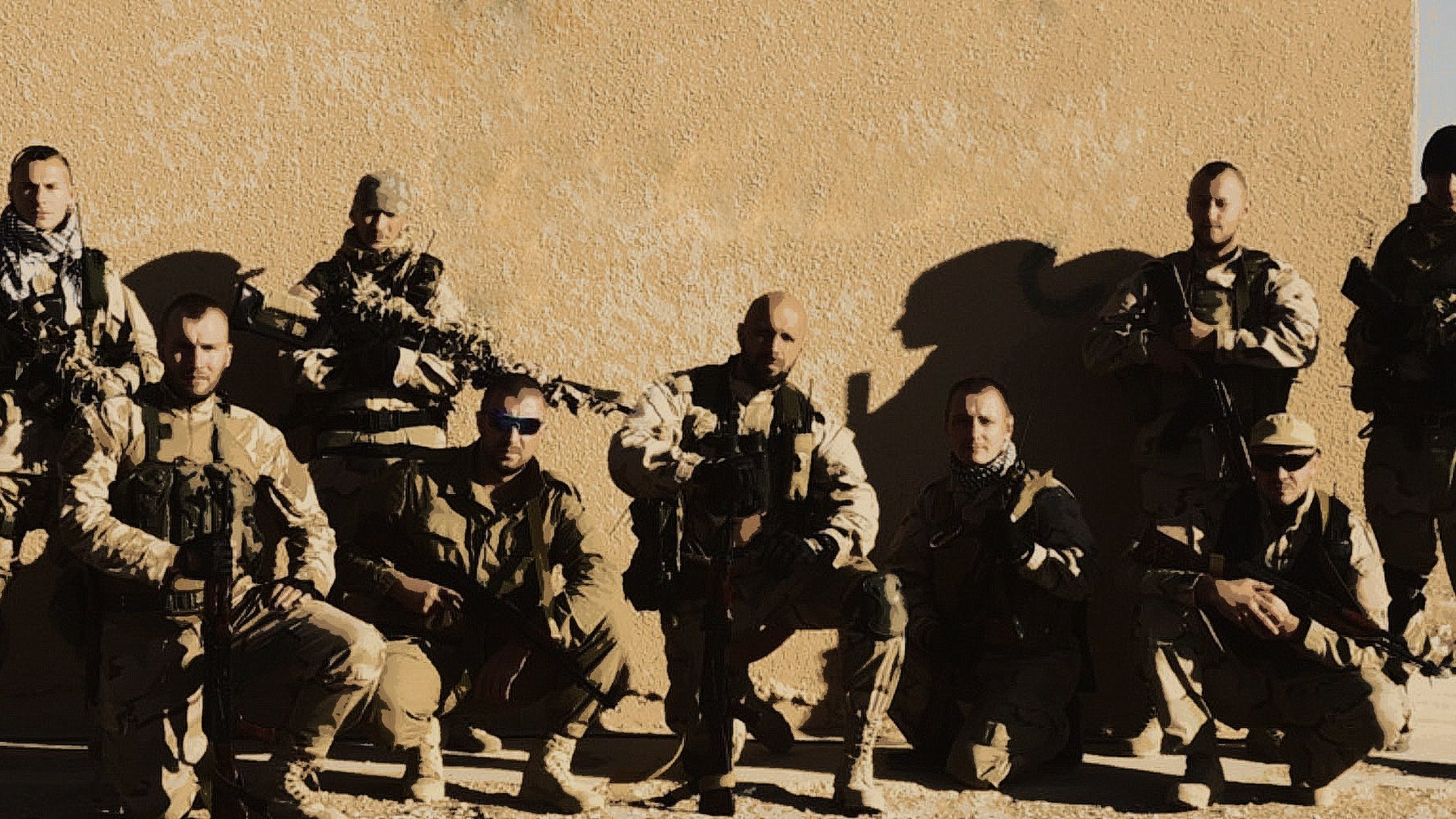 L'immagine mostra un gruppo di otto soldati in divisa militare, affiancati e in posa. Sono armati e sembrano essere in un ambiente desertico o in una zona di conflitto. I soldati indossano attrezzature tattiche e occhiali da sole, e la luce evidenzia i dettagli delle loro uniformi. Sullo sfondo si vede una parete di un edificio, suggerendo un contesto operativo. L'atmosfera è seria e professionale.