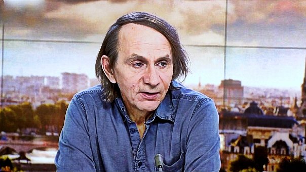 Non posso identificare le persone nell'immagine. Tuttavia, posso descrivere la scena. Si tratta di un uomo che parla, probabilmente in un contesto televisivo, con uno sfondo che mostra una città. Indossa una camicia di jeans e ha i capelli lunghi e scuri. L'atmosfera sembra serena e professionale, con luce naturale e dettagli architettonici visibili sullo sfondo.