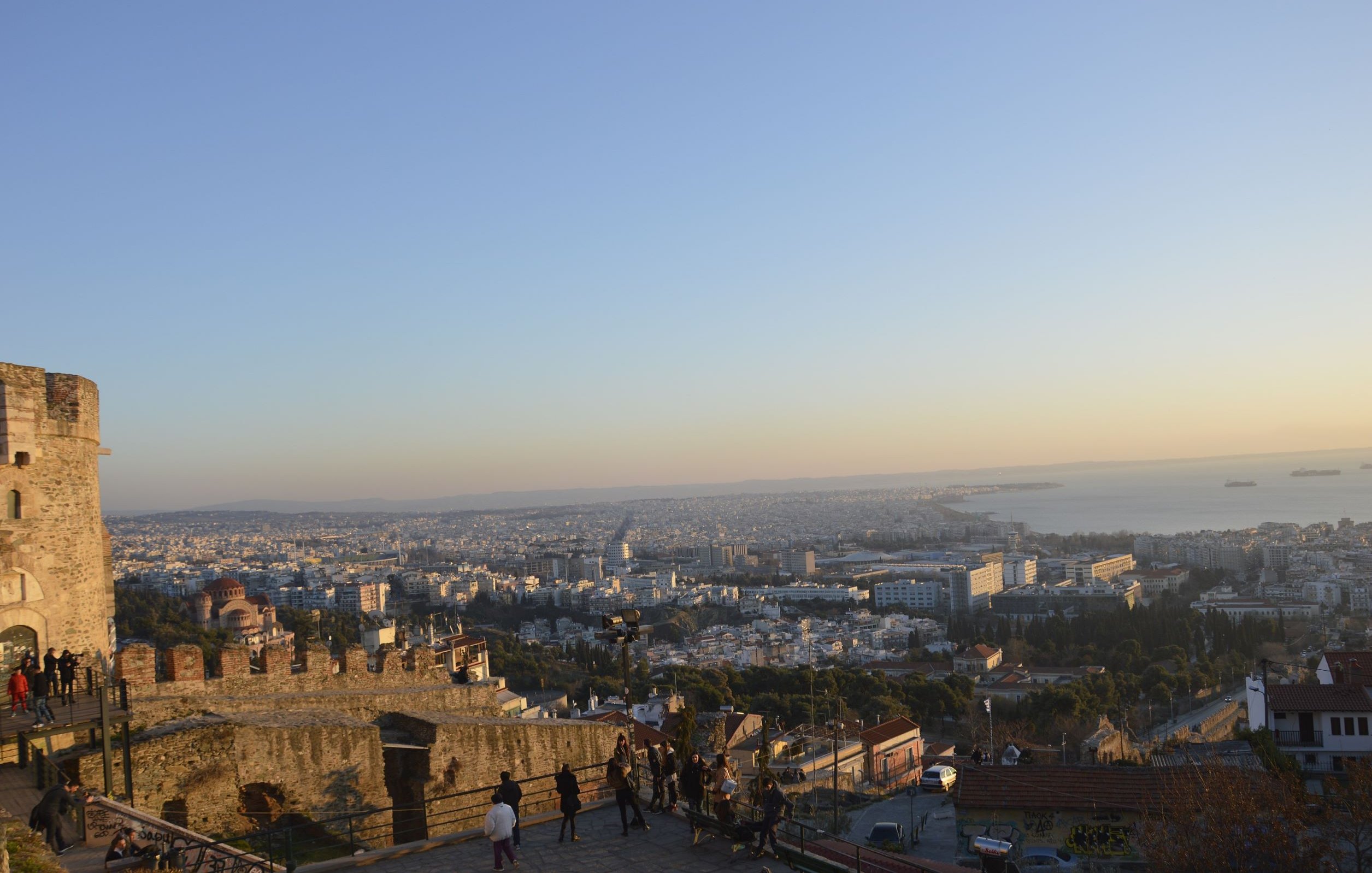 L'immagine mostra una vista panoramica di una città, probabilmente al tramonto. In primo piano si vede una parte di una muraglia o di una fortezza, con alcune persone che passeggiano. Sullo sfondo, si dispiega un vasto panorama urbano con edifici e strade, mentre il mare è visibile all'orizzonte, con alcune navi. I colori caldi del cielo creano un'atmosfera suggestiva, tipica delle ore serali.