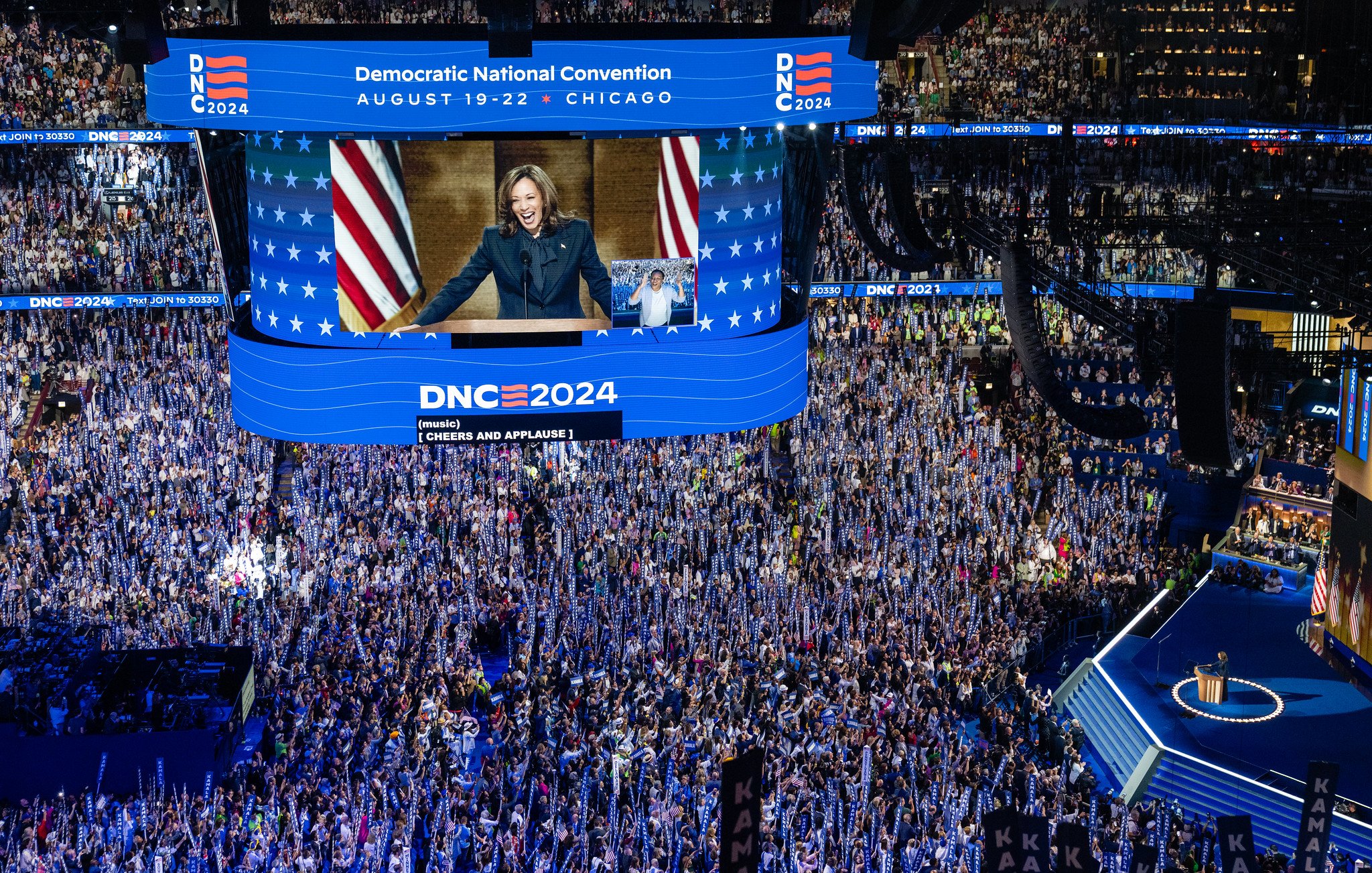 L'immagine mostra una scena di una grande convention politica, probabilmente la Convenzione Nazionale Democratica del 2024 a Chicago. La sala è affollata di persone che applaudono, con molti sostenitori che sventolano bandiere e cartelli. Al centro, un grande schermo mostra un discorso di una figura politica (probabilmente un leader del partito). Gli ambienti sono decorati con colori e simboli del partito, come stelle e strisce. L'atmosfera sembra essere di entusiasmo e impegno politico.