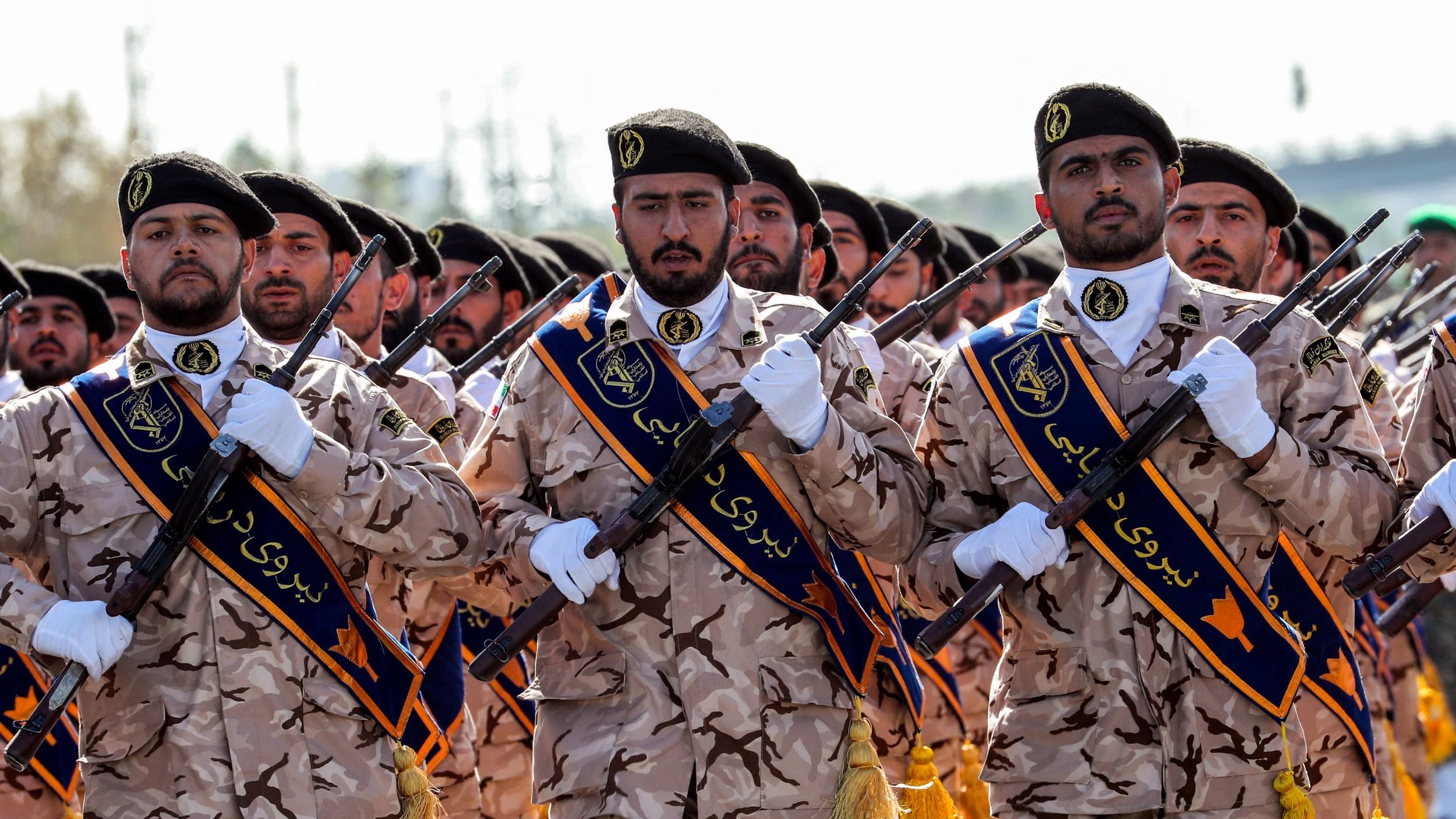 L'immagine mostra un gruppo di uomini in uniforme che sfilano. Indossano abiti mimetici e berretto, con fasce distintive sui loro uniformi. Alcuni di loro tengono in mano fucili, tutti sembrano professionali e seri. Lo sfondo è sfocato, ma suggerisce un luogo di parata o celebrazione ufficiale. L'atmosfera sembra essere di solennità e disciplina.