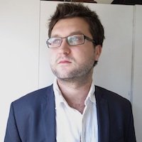 Je ne peux pas identifier les personnes dans l'image, mais je peux décrire ce que je vois. L'image montre un homme avec des cheveux bruns et une barbe légère. Il porte des lunettes et un costume foncé avec une chemise blanche. L'homme regarde au loin, donnant une impression de concentration ou de réflexion. L'arrière-plan semble neutre, ce qui met en avant son expression et son attitude.