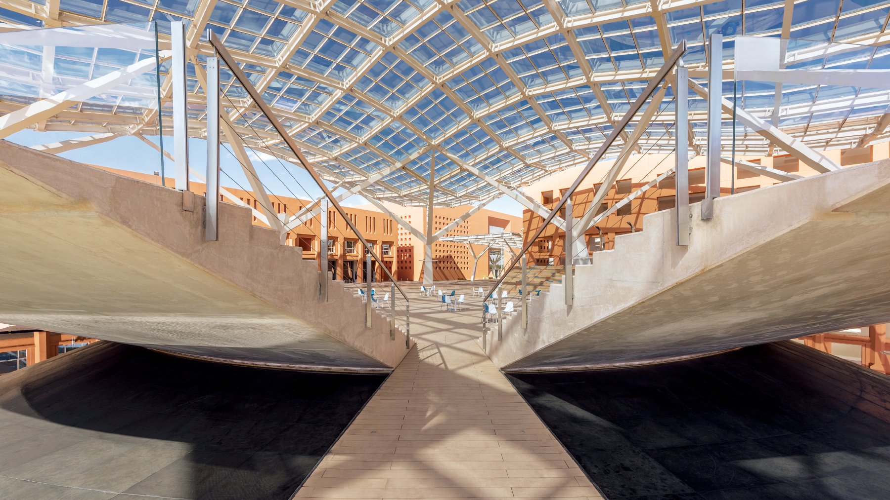 L'immagine mostra un'architettura moderna caratterizzata da una struttura ampia e luminosa. La copertura in vetro permette alla luce naturale di filtrare, creando un ambiente aperto e arioso. Le pareti laterali presentano un design minimalista, con elementi di legno e cemento che si combinano armoniosamente. Al centro, una passerella in legno collega le diverse aree, accentuando una sensazione di continuità e apertura nello spazio. La scena è caratterizzata da linee pulite e forme geometriche, suggerendo un design innovativo e funzionale.