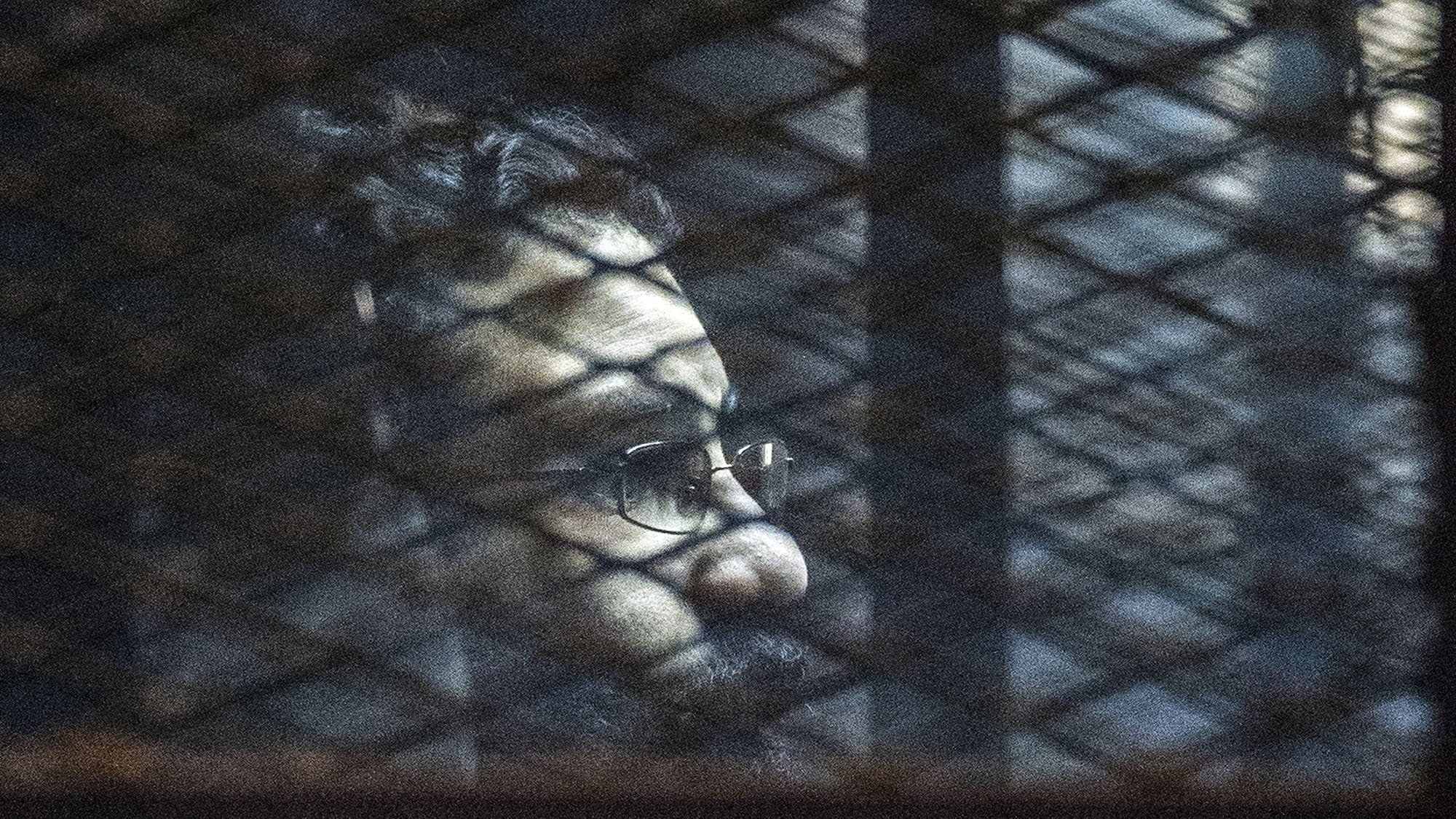 L'immagine mostra un uomo visto attraverso una rete metallica. La luce crea ombre sul suo viso, evidenziando la sua espressione seria. Indossa occhiali e ha una barba, mentre lo sfondo risulta scuro e indistinto, contribuendo a un'atmosfera di introspezione o solitudine.