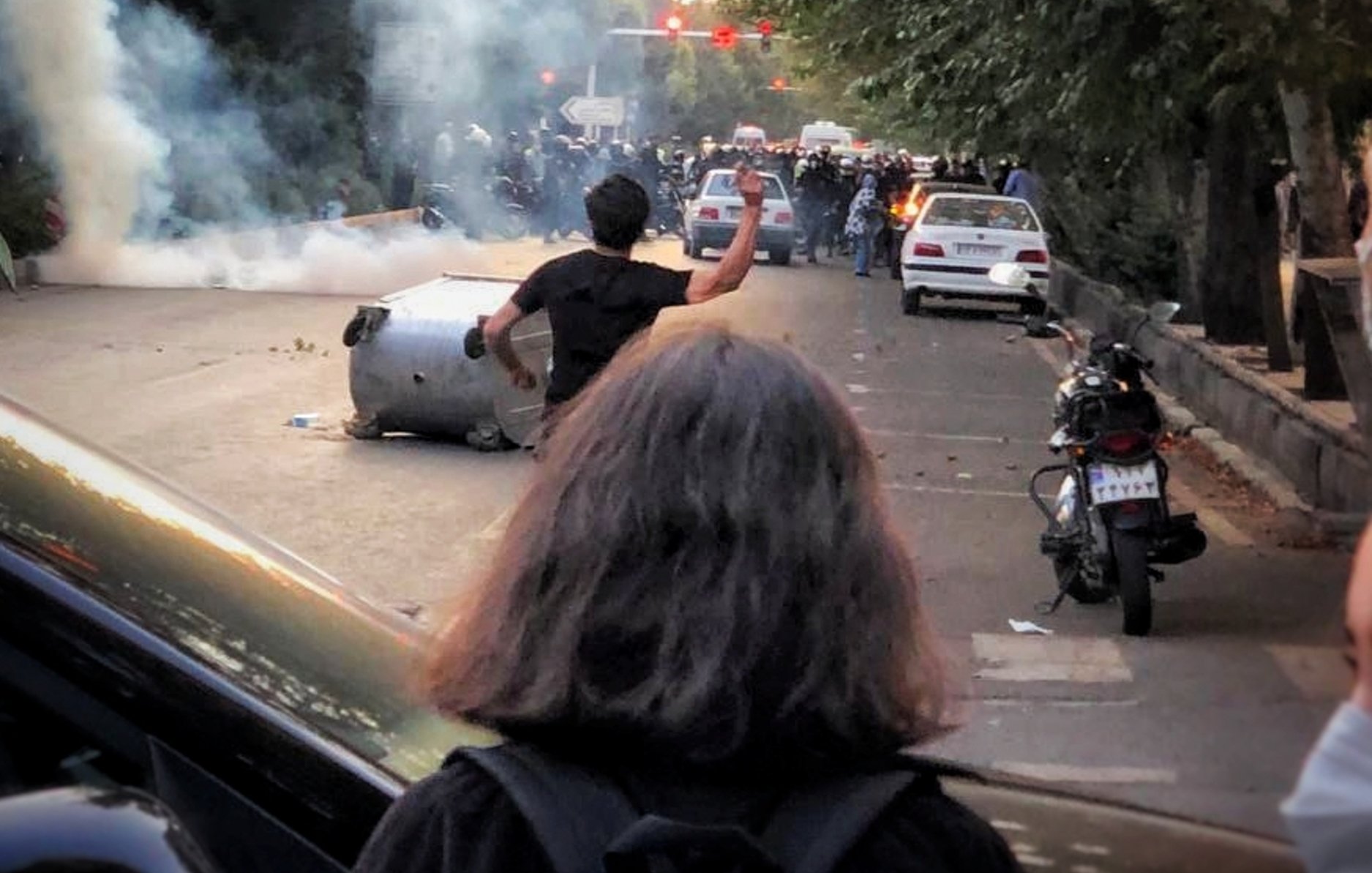 L'immagine mostra una scena di tensione e conflitto in una strada. Ci sono persone in piedi, alcune sembrano protestare, e si nota un'atmosfera di caos con fumi o gas lacrimogeni nell'aria. Alcuni veicoli sono coinvolti, con una macchina ribaltata, mentre altri autoveicoli sono parcheggiati lungo il marciapiede. Gli alberi ai lati della strada contribuiscono a dare un'idea di una zona urbana. La persona in primo piano, che ha i capelli lunghi, sembra stia alzando un braccio verso qualcosa, forse in segno di protesta o sfida.