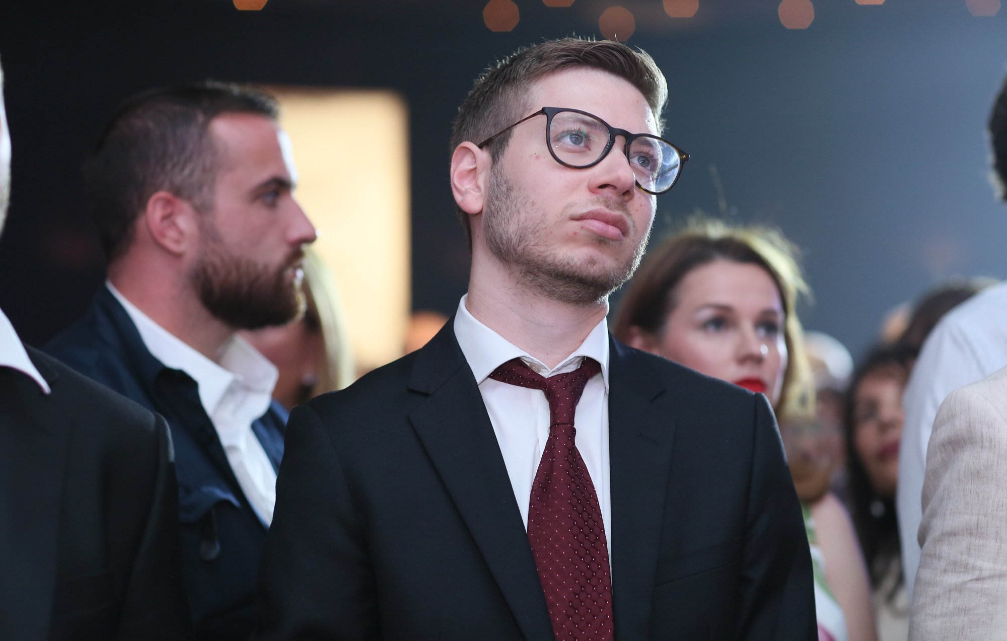 Nell'immagine si può vedere un gruppo di persone in un contesto che sembra essere un evento sociale o una festa. Un uomo in primo piano indossa un completo scuro con una cravatta bordeaux e occhiali. Ha un'espressione pensierosa, mentre osserva attentamente ciò che accade di fronte a lui. Dietro di lui ci sono altre persone, alcune delle quali sembrano anch'esse concentrate sull'eventuale spettacolo o presentazione. L'atmosfera è elegante e formale, con una leggera illuminazione che aggiunge un tocco di atmosfera.