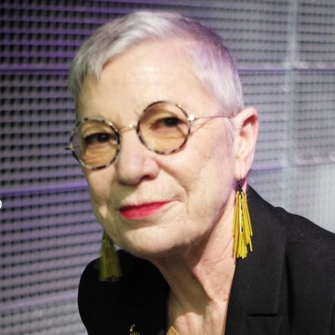 L'image montre une femme d'un certain âge avec des cheveux courts et gris. Elle porte des lunettes rondes et de grandes boucles d'oreilles jaunes. Son expression est sérieuse et elle semble regarder vers l'objectif. Elle est habillée en noir, ce qui contraste avec son teint clair. L'arrière-plan est flou et paraît moderne.