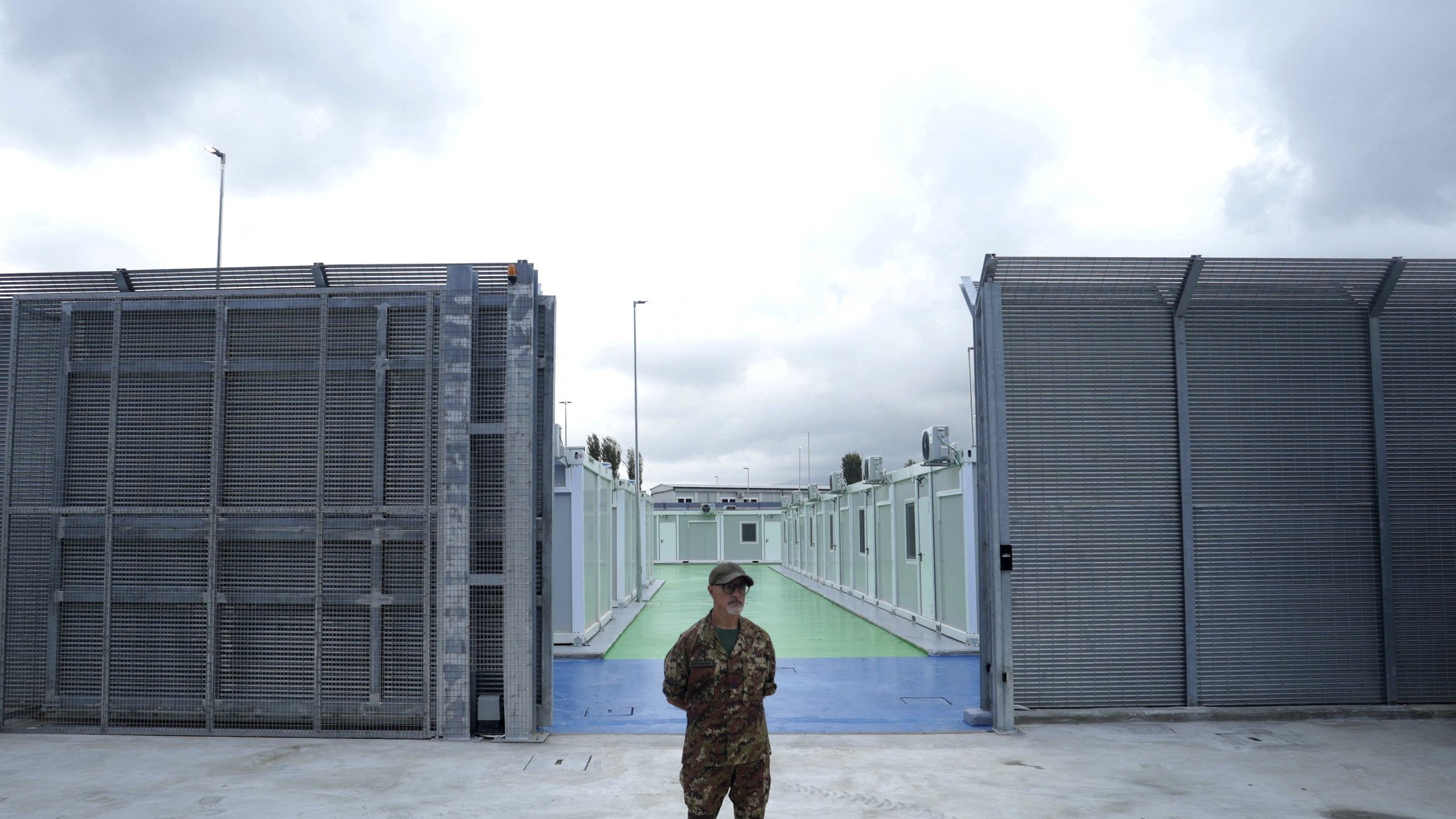 L'immagine mostra un soldato in uniforme, in piedi al centro di un'area delimitata. Sullo sfondo si possono vedere due strutture metalliche grigie che sembrano essere recinzioni, con un'area di pavimentazione colorata di verde e blu tra di esse. Il cielo è nuvoloso, suggerendo una giornata grigia. L'atmosfera sembra essere di sicurezza e controllo, con un forte senso di vigilanza da parte del soldato.