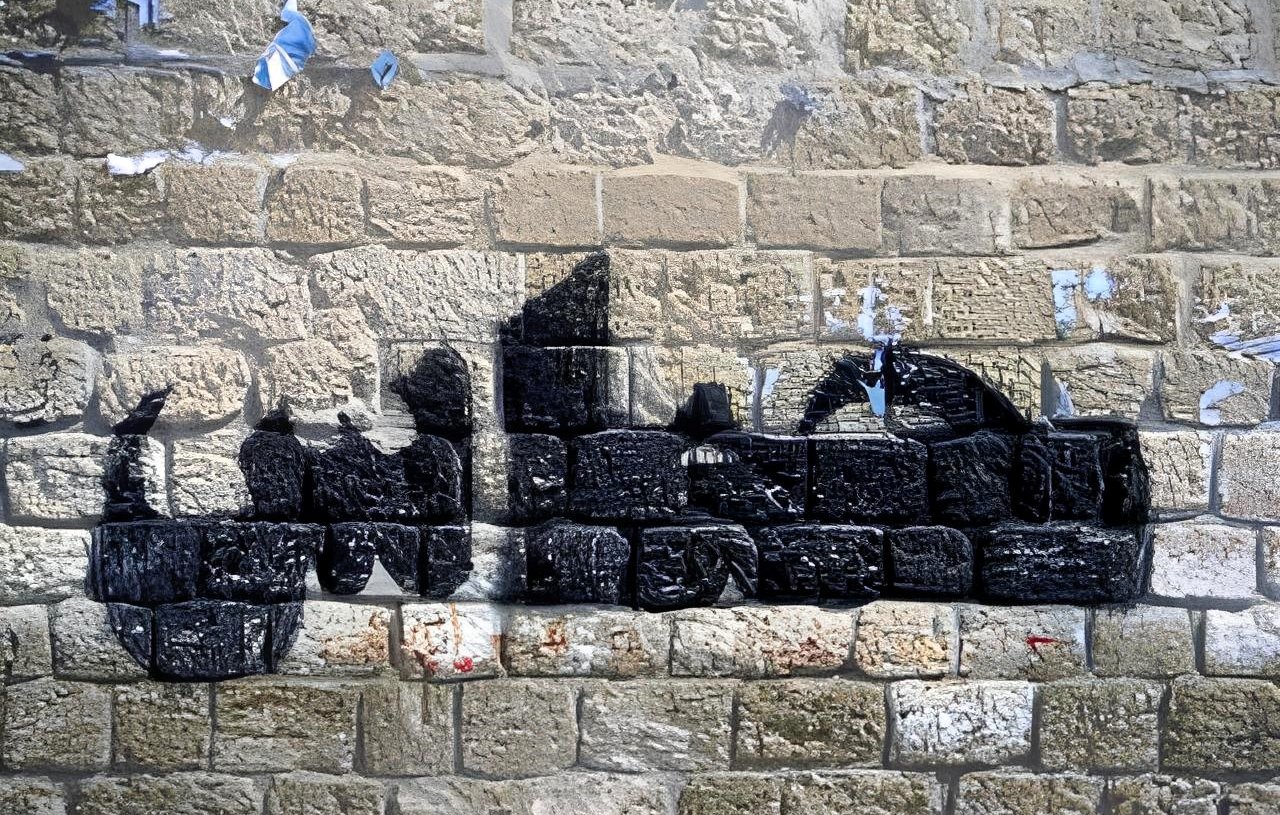 L'immagine mostra un murale su un muro di pietra. Il disegno rappresenta una silhouette di un veicolo, probabilmente un camion, realizzato con vernice nera. Le linee e le forme sono stilizzate, creando un contrasto con la texture della parete di pietra. Sul muro si notano anche resti di manifesti strappati e segni di usura, che aggiungono carattere e storia all'ambiente.