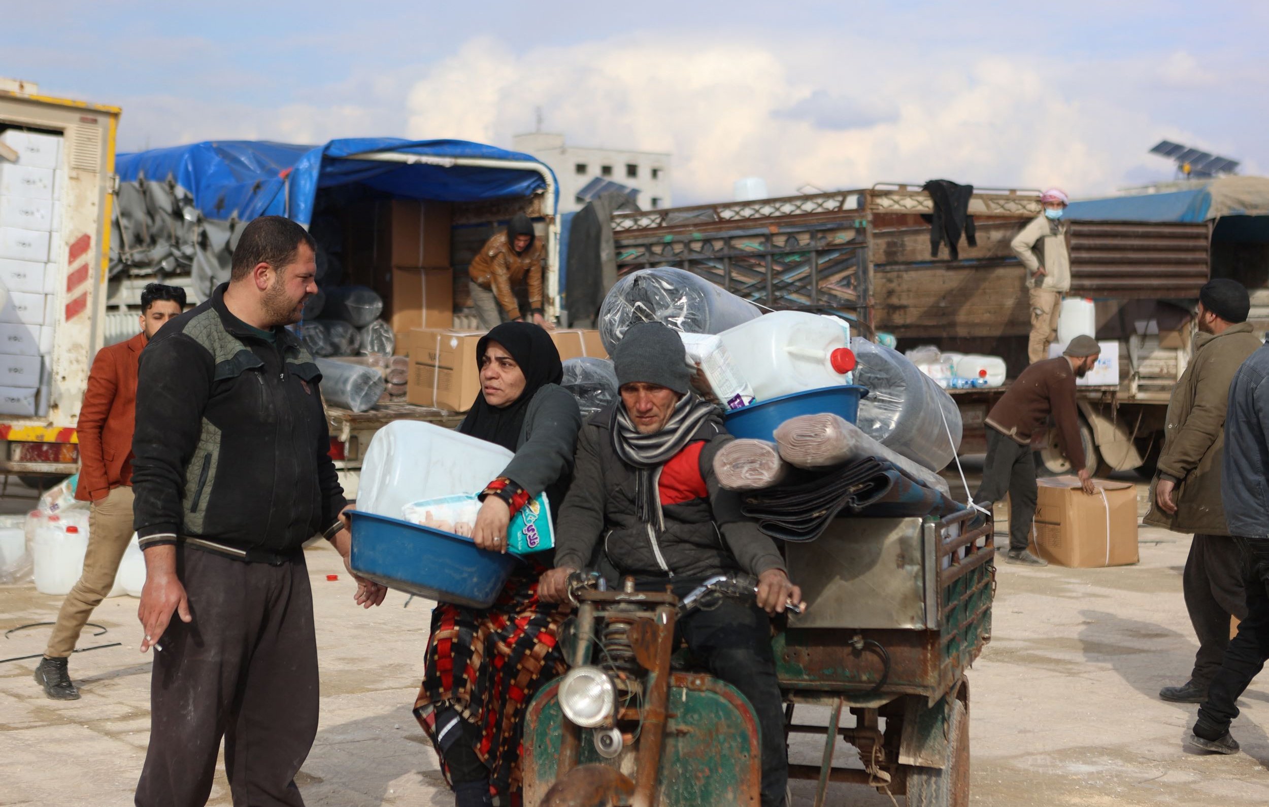 Nell'immagine si vede una scena di un'area di stoccaggio o mercato. Ci sono alcune persone che sembrano occupate a scaricare e trasportare materiali. Un uomo e una donna sono in primo piano: l'uomo indossa abiti scuri e tiene una cassetta, mentre la donna è vestita con un lungo mantello scuro e apparente una certa preoccupazione sul volto. Dietro di loro, ci sono merci come bottiglie di plastica e tappeti, oltre a veicoli e altri uomini che svolgono attività di carico e scarico. L'atmosfera sembra essere quella di un'operazione di supporto umanitario.