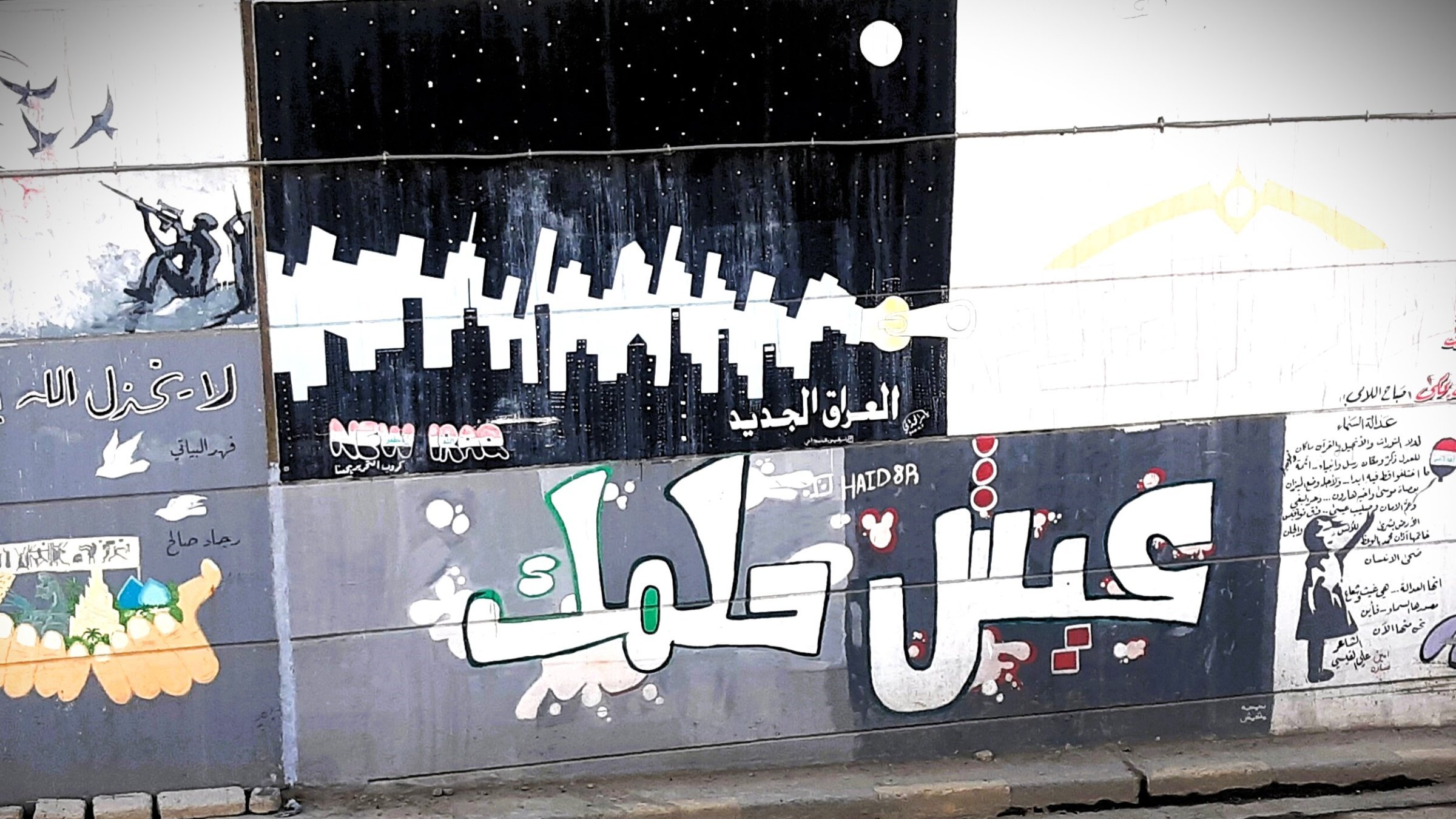 L'immagine mostra un murale colorato su un muro, con diversi disegni e scritte in arabo. In primo piano, si legge "عيش حلمك" che significa "Vivi il tuo sogno". Lo sfondo presenta vari elementi artistici, tra cui silhouette di grattacieli e figure stilizzate, creando un'atmosfera urbana e vibrante. Ci sono anche elementi decorativi e simboli che riflettono un messaggio di speranza e ispirazione.