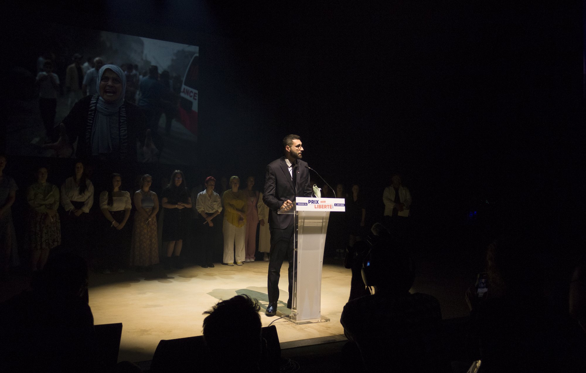 L'immagine mostra un uomo che parla al microfono su un palco, mentre sullo sfondo si vede un'immagine proiettata di una donna con un velo e altre persone. L'oratore indossa un abito elegante e sembra essere il centro dell'attenzione. Dietro di lui, altre persone sono schierate, probabilmente in segno di supporto o partecipazione all'evento. L'illuminazione è drammatica, creando un'atmosfera intensa e concentrata sull'oratore.