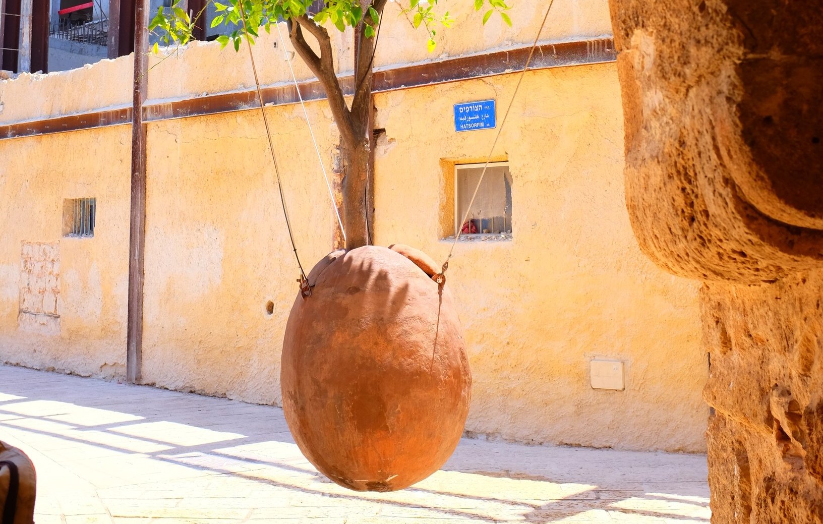 L'immagine mostra un quartiere caratteristico con un muro di colore giallo chiaro. Al centro, si vede un grande vaso di terra cotta che oscilla, sospeso con delle corde. Il vaso sembra appeso a un albero, mentre sullo sfondo si intravede un edificio con strutture in legno e finestre. La scena appare luminosa e vivace, con una sensazione di atmosfera calda e accogliente.