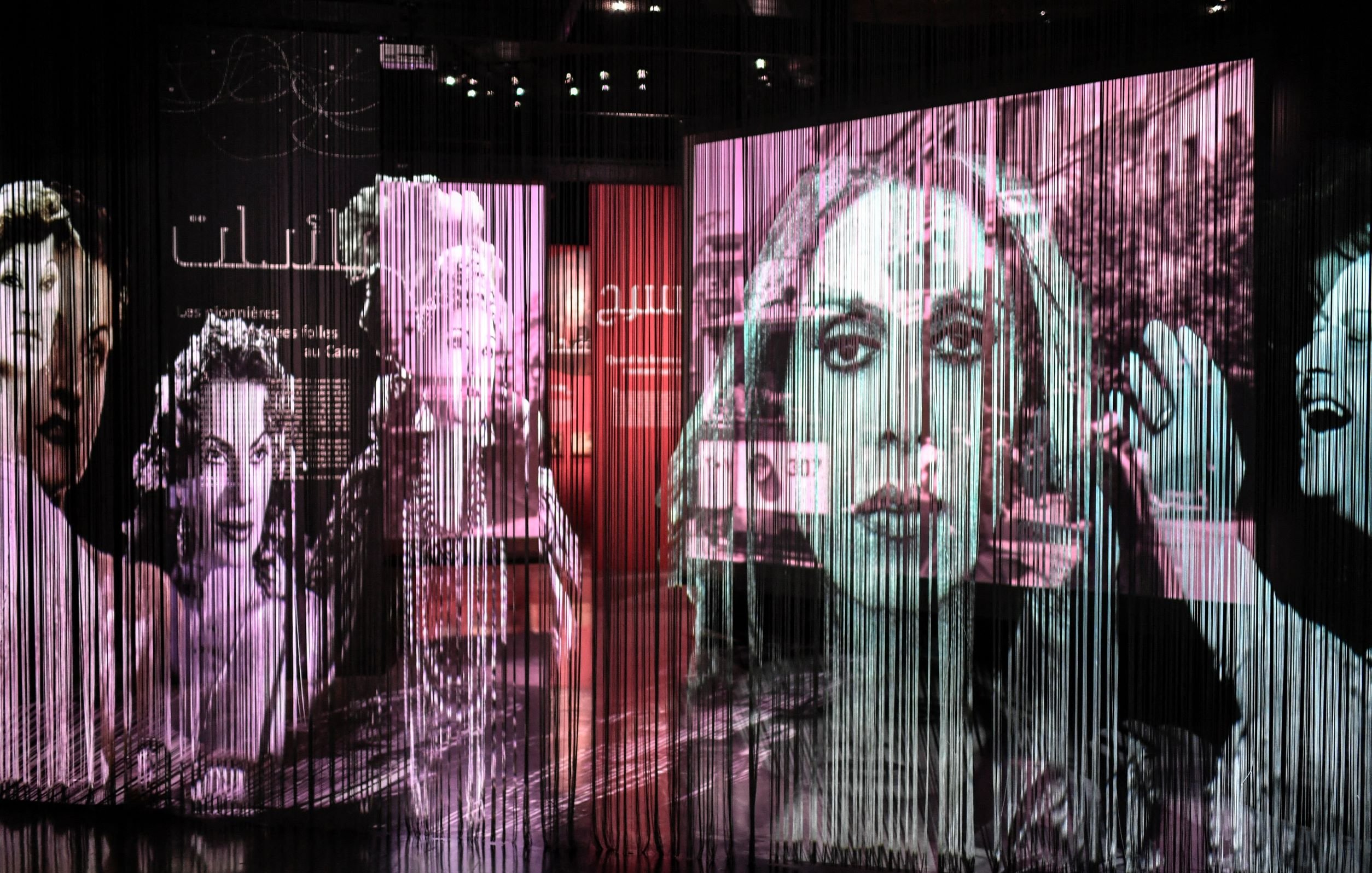 L'immagine presenta una serie di proiezioni artistiche su uno sfondo di tende di tessuto trasparente. Le immagini ritraggono volti femminili e scene di vita, con un forte contrasto tra bianco e nero e tonalità colorate. L'atmosfera è suggestiva e intimista, creando un effetto di profondità e movimento. Le fotografie sembrano evocare un senso di nostalgia, magari legato alla storia e alla cultura. Le scritte in arabo e altre lingue suggeriscono un tema culturale e un messaggio profondo.