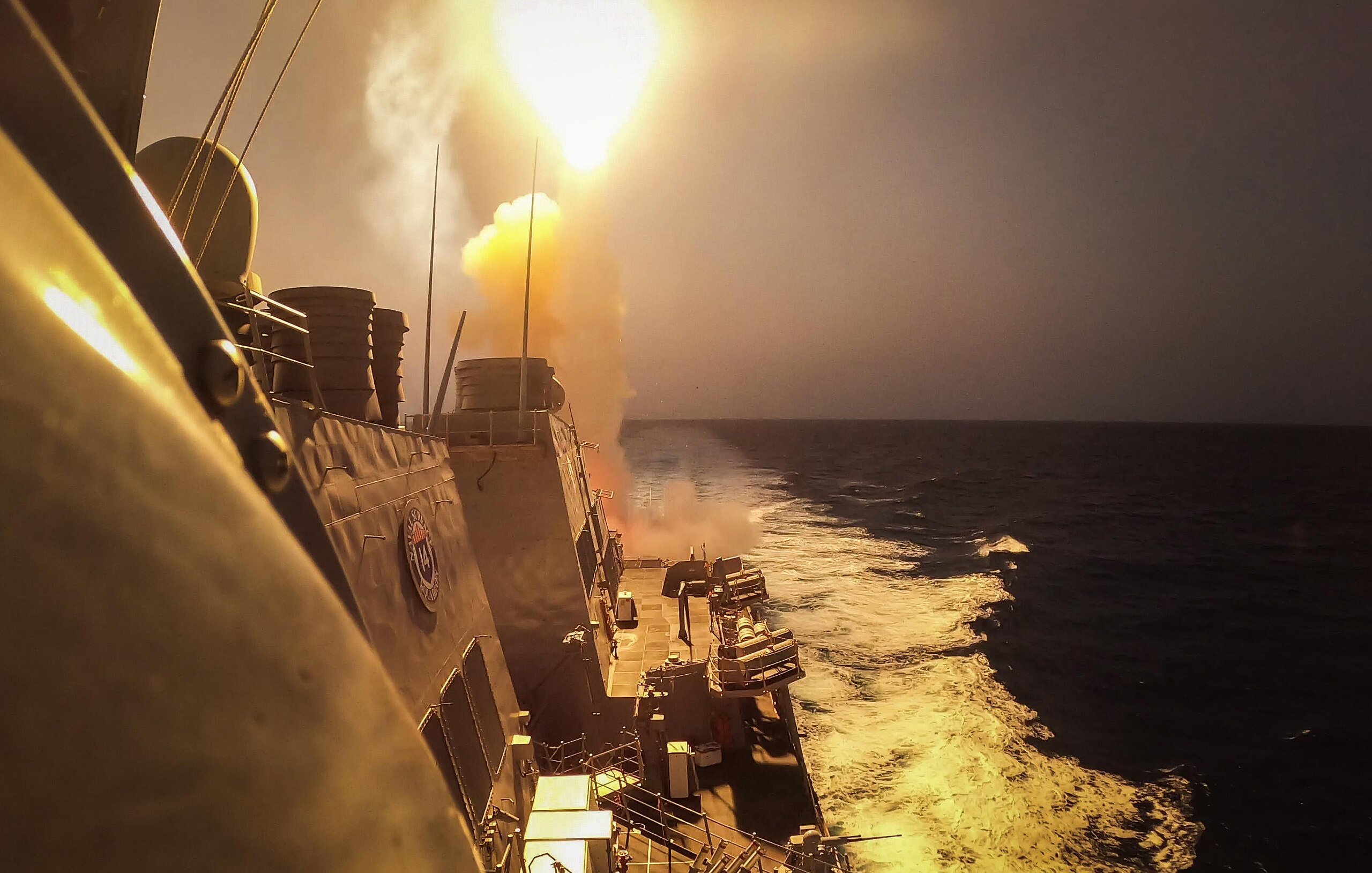 L'immagine mostra una scena marittima in cui un missile viene lanciato da una nave da guerra. Si vede il missile che viene sparato con un forte bagliore e un'esplosione di fumi dietro di esso. In primo piano si possono notare le strutture della nave e il mare in movimento, evidenziato da schiuma bianca. Il cielo appare velato e si intuisce un momento di grande intensità e potenza militare.