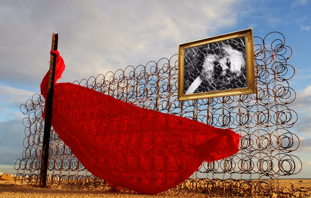 L'immagine mostra una scena poetica e surreale. Al centro, un drappo rosso fluttua nel vento, appendendosi a una struttura metallica composta da spirali di ferro. Dietro il drappo, vi è una cornice dorata che contiene una fotografia in bianco e nero, probabilmente di un soggetto significativo. Il cielo sullo sfondo è ampio, con nuvole sparse che creano un'atmosfera suggestiva. L'insieme evoca temi di libertà, memoria e contrasto tra materiali ruvidi e il delicato tessuto rosso.