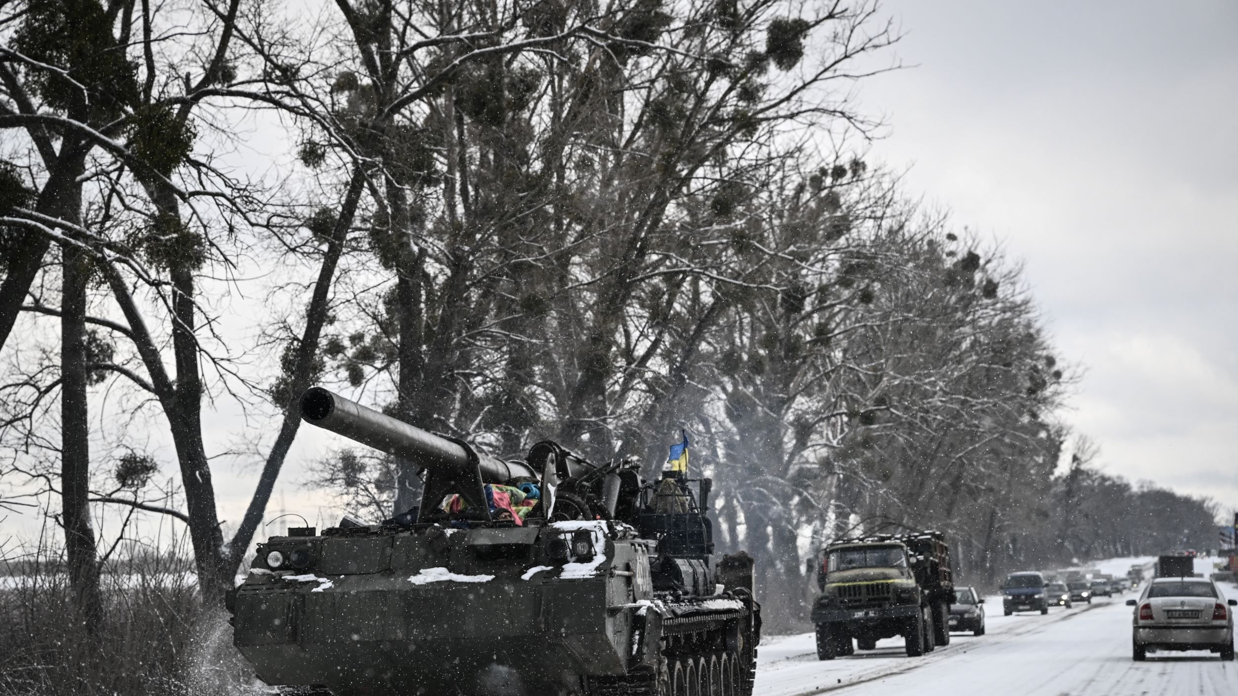 L'immagine mostra un carro armato che si muove lungo una strada innevata, circondato da alberi. Il carro armato ha una canna protrudente e presenta dettagli decorativi che potrebbero indicare l'appartenenza a una specifica unità militare. Accanto al carro armato, si intravedono anche alcuni veicoli, probabilmente militari, che viaggiano sulla stessa strada. Il paesaggio è caratterizzato da un'atmosfera invernale, con la neve che copre sia la strada che il terreno circostante.
