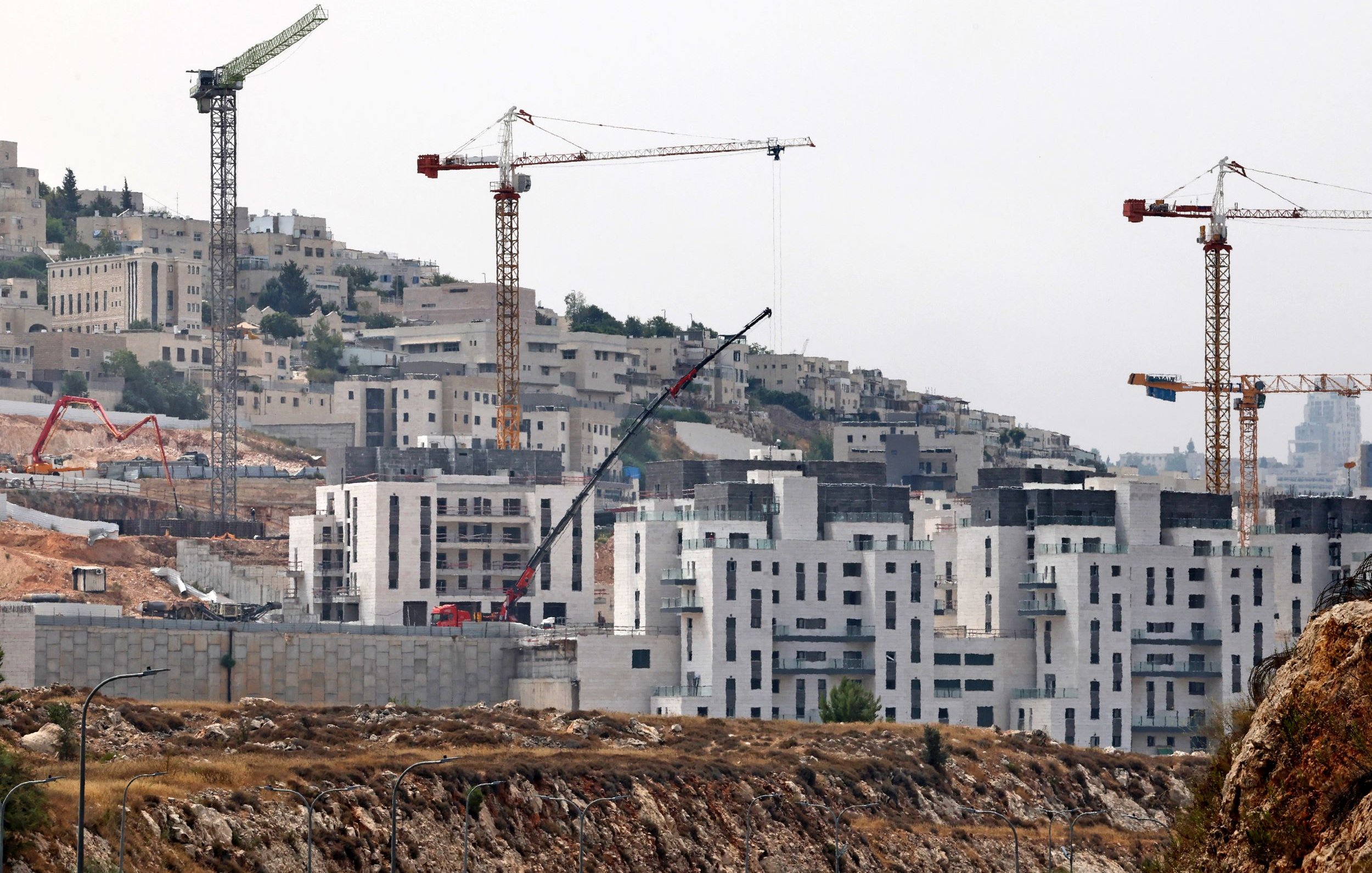 L'immagine mostra un cantiere edile situato in una zona collinare. Sono visibili diverse gru da costruzione, che si ergono sopra gli edifici in fase di completamento. Le strutture in costruzione hanno una forma moderna e presentano superfici di cemento. Sullo sfondo si intravedono altre costruzioni e il panorama collinare circostante, con vegetazione sparsa. L'atmosfera sembra essere quella di un'area in sviluppo e crescita urbana.