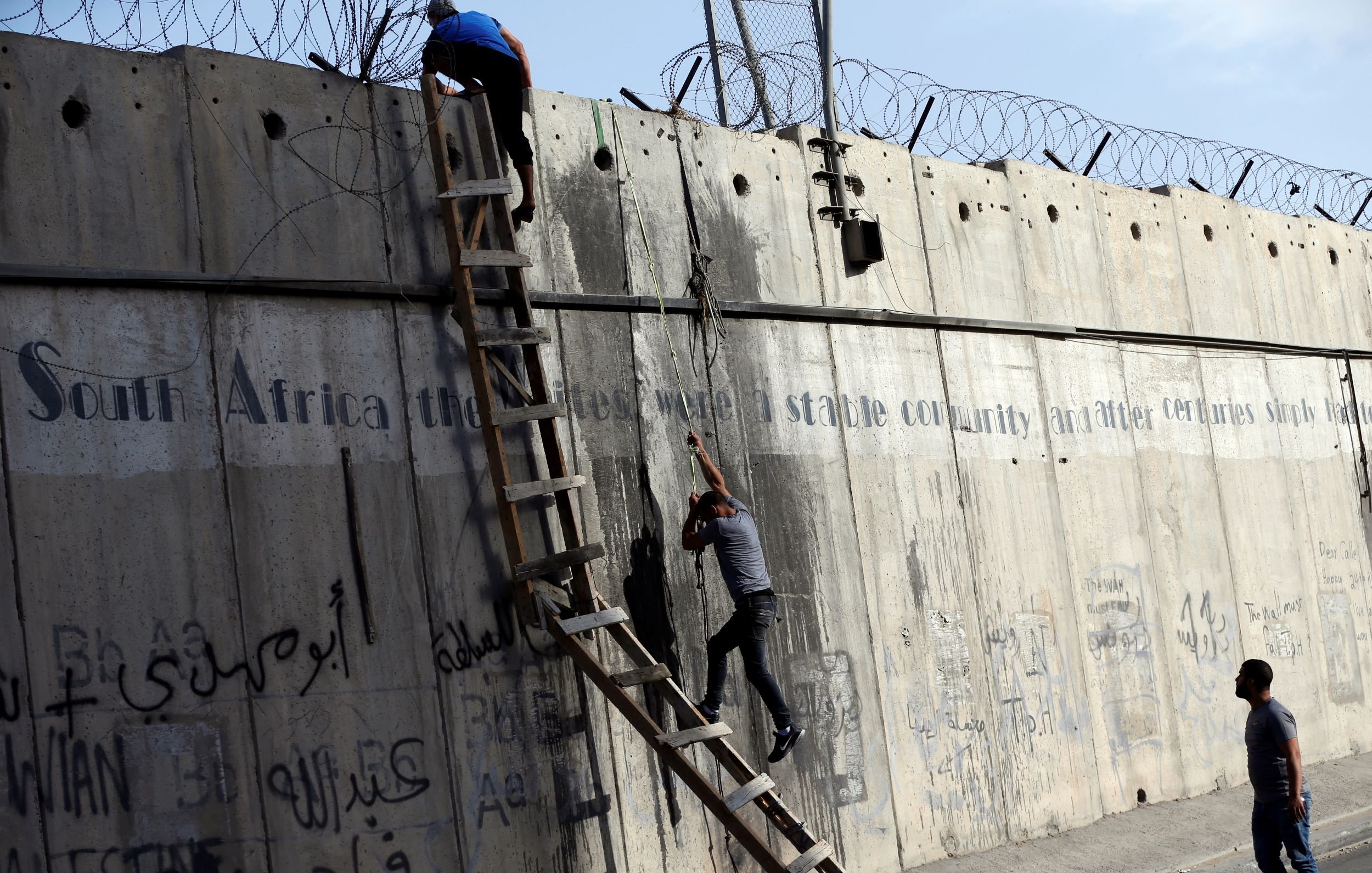 L'immagine mostra due persone che stanno scalando un muro di cemento, sul quale è presente della rete metallica. Dietro di loro, è visibile una scala che qualcuno sta utilizzando per salire. Il muro è coperto da graffiti e scritte, in varie lingue, che sembrano affrontare temi legati alla storia e alla condizione sociale. La scena evoca un senso di tensione e disperazione, con le persone che tentano di superare un ostacolo fisico, simbolo di divisione.