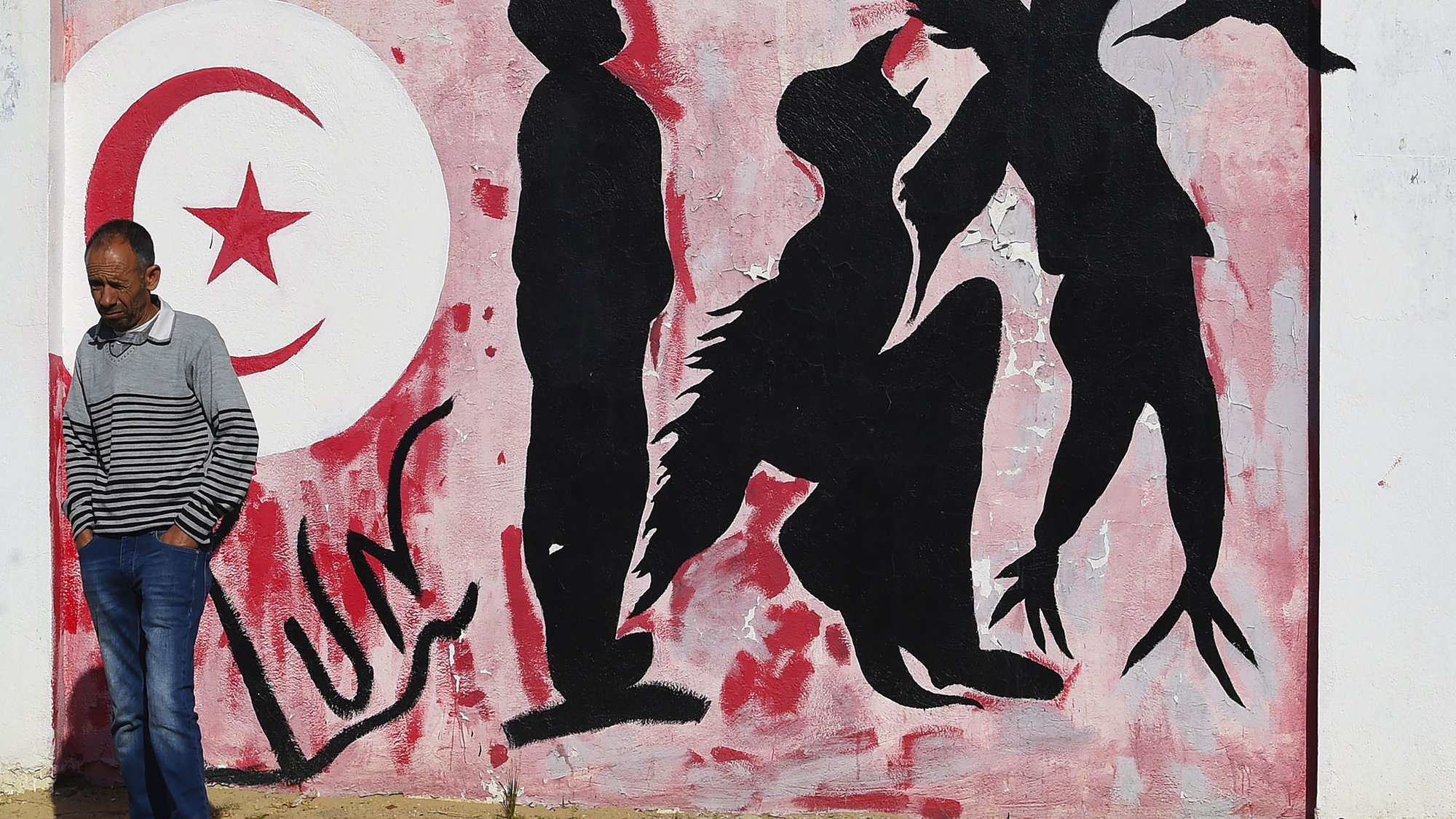 L'immagine mostra un murale su un muro bianco, caratterizzato da una palette di colori rossi e neri. Sullo sfondo, è presente un grande simbolo della luna crescente e una stella, simboli spesso associati alla bandiera tunisina. Al centro, ci sono delle silhouette di figure umane che appaiono in diverse posizioni: alcune sono in piedi, altre sembrano inginocchiarsi o muoversi in modo dinamico. In primo piano, un uomo con una maglietta a strisce passeggia accanto al murale, creando un contrasto tra l'arte e la vita quotidiana. L'atmosfera trasmette un senso di cultura e identità, evidentemente legato alla Tunisia.
