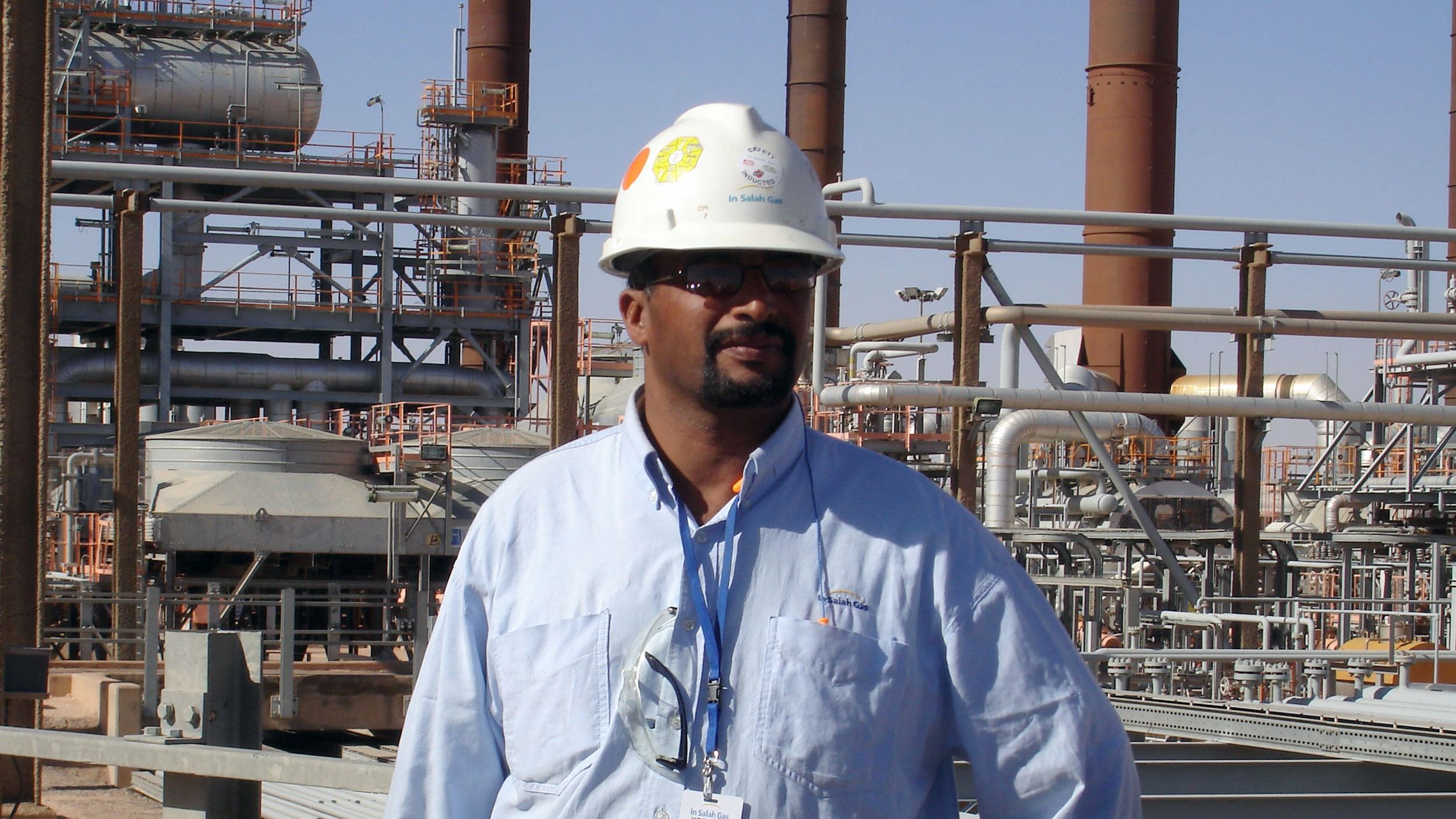 L'immagine mostra un uomo che indossa un casco di sicurezza bianco e una camicia azzurra. Si trova in un ambiente industriale, con impianti e tubazioni sullo sfondo. L'atmosfera sembra quella di un sito di produzione o di una raffineria, con diverse strutture metalliche e torri visibili. L'uomo appare fiducioso e professionale mentre si pone di fronte all'obiettivo.