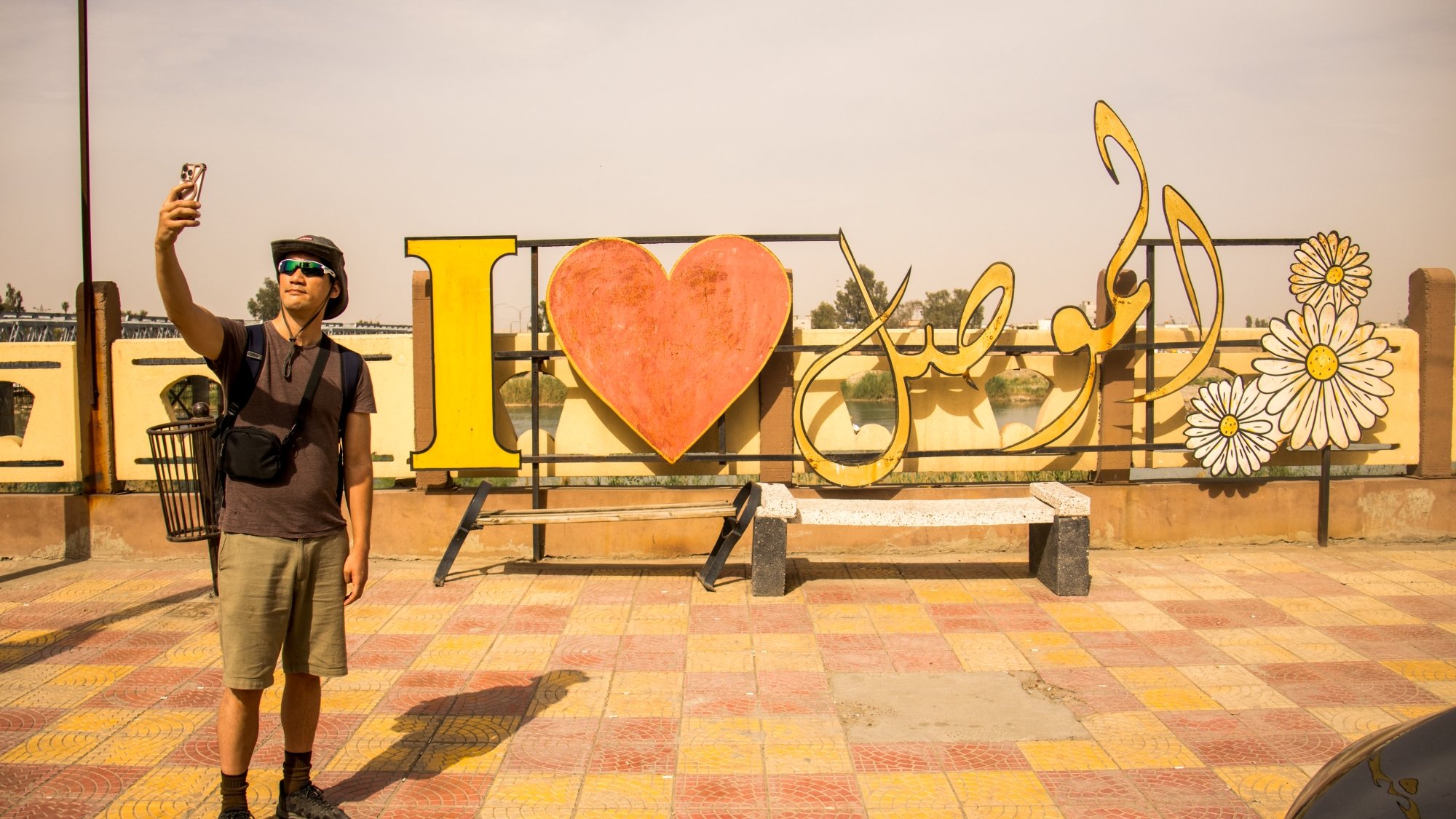 Un uomo fa un selfie davanti a un grande cartello colorato con "I ❤️" e fiori.
