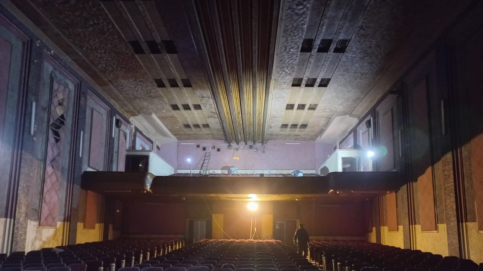 L'immagine mostra l'interno di un teatro abbandonato. Si possono vedere file di poltrone di velluto viola, di fronte a un palcoscenico che sembra vuoto. La luce proveniente da un proiettore evidenzia il pavimento e crea un'atmosfera suggestiva. Sul palcoscenico, ci sono alcune attrezzature, come scale, suggerendo che ci siano lavori in corso o recenti ispezioni. Le pareti del teatro mostrano segni di degrado, contribuendo a un senso di malinconia e nostalgia.