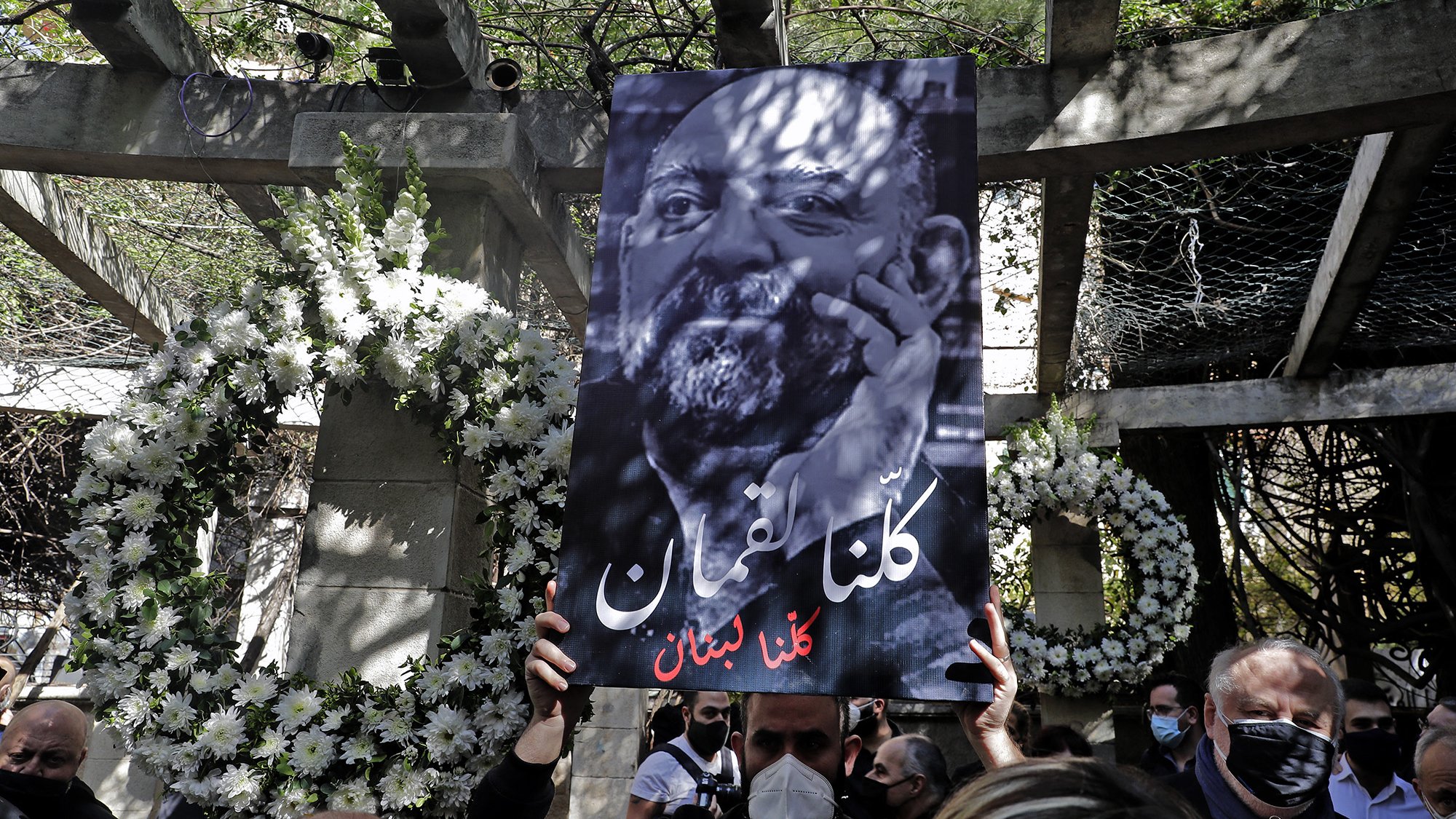 L'immagine mostra una persona che tiene in alto un grande cartello. Il cartello presenta un ritratto in bianco e nero di un uomo con una barba, accompagnato da un testo in arabo che significa "Siamo tutti Libano". Sullo sfondo, ci sono decorazioni floreali e un ambiente che sembra essere un luogo commemorativo. La scena trasmette un senso di unità e solidarietà.