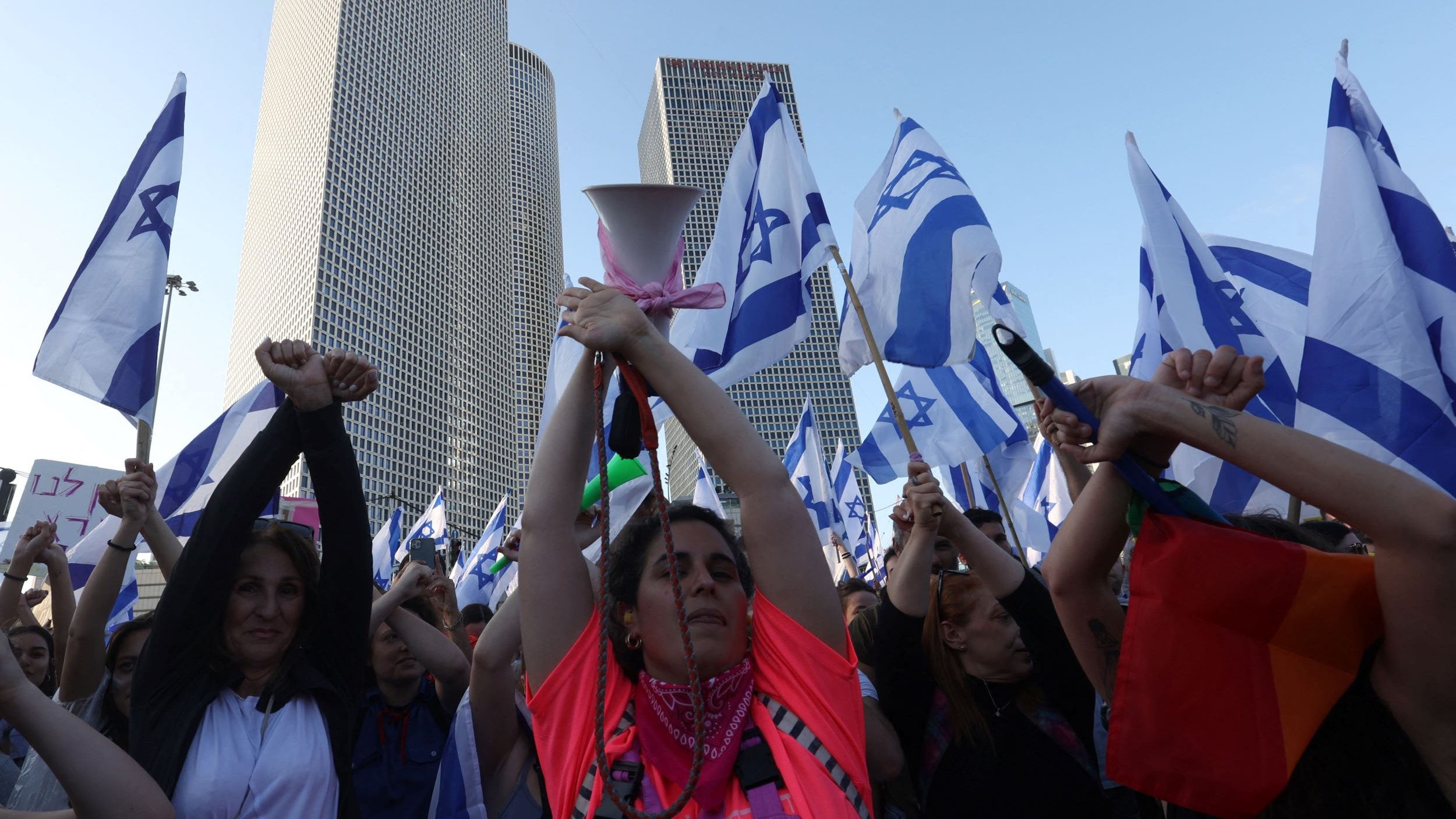 L'immagine mostra una folla di persone che partecipa a una manifestazione. Alcuni partecipanti tengono in alto delle bandiere israeliane, mentre molti altri alzano le mani, dimostrando un senso di unità e protesta. In primo piano, si può vedere una donna con una maglietta di colore neon che sembra guidare la manifestazione con un megafono. Sullo sfondo, si notano alti edifici moderni, suggerendo che l'evento si svolge in un contesto urbano. L'atmosfera è carica di energia e determinazione.
