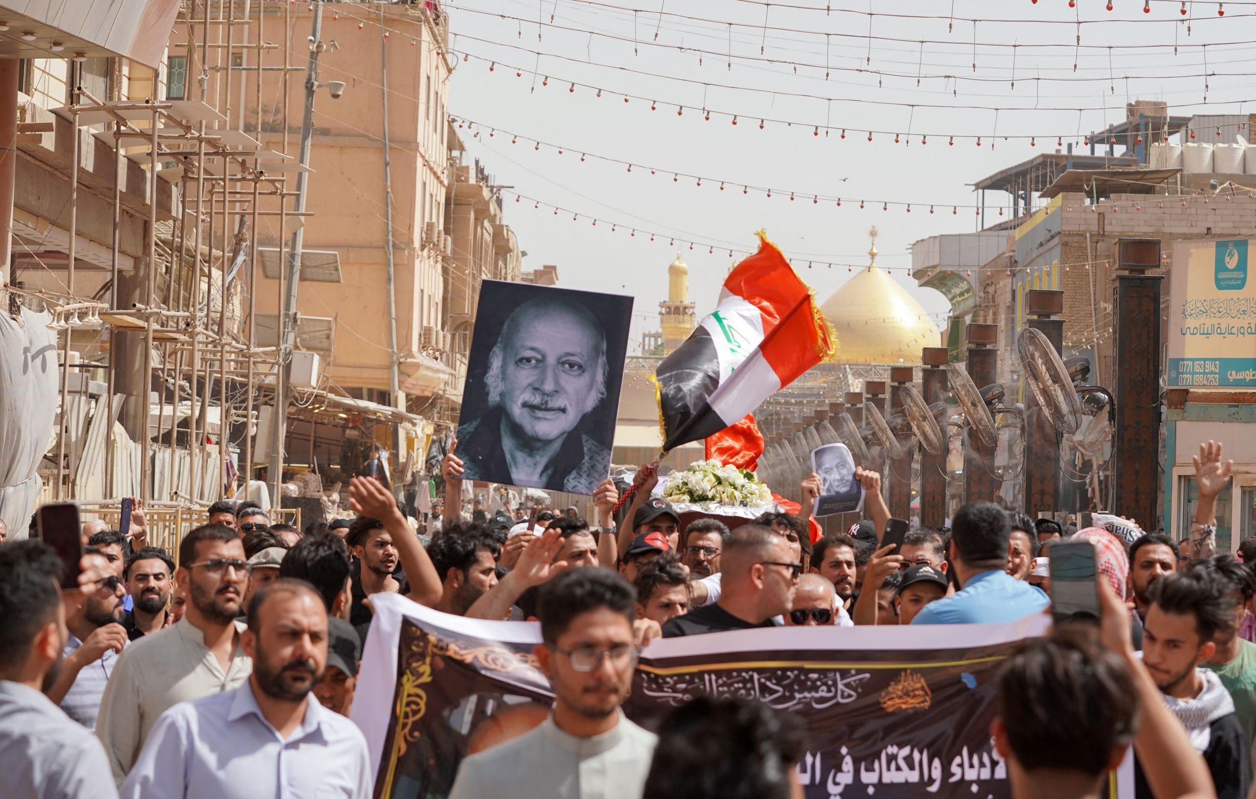 L'immagine mostra una folla di persone durante una manifestazione. Molti manifestanti tengono in alto una grande fotografia di un uomo, insieme a una bandiera irachena. Sullo sfondo si possono vedere elementi architettonici e due cupole dorate, che suggeriscono la presenza di un luogo sacro o importante. L'atmosfera sembra intensa e significativa, con i partecipanti che mostrano il loro sostegno o commemorazione attraverso slogan e simboli.