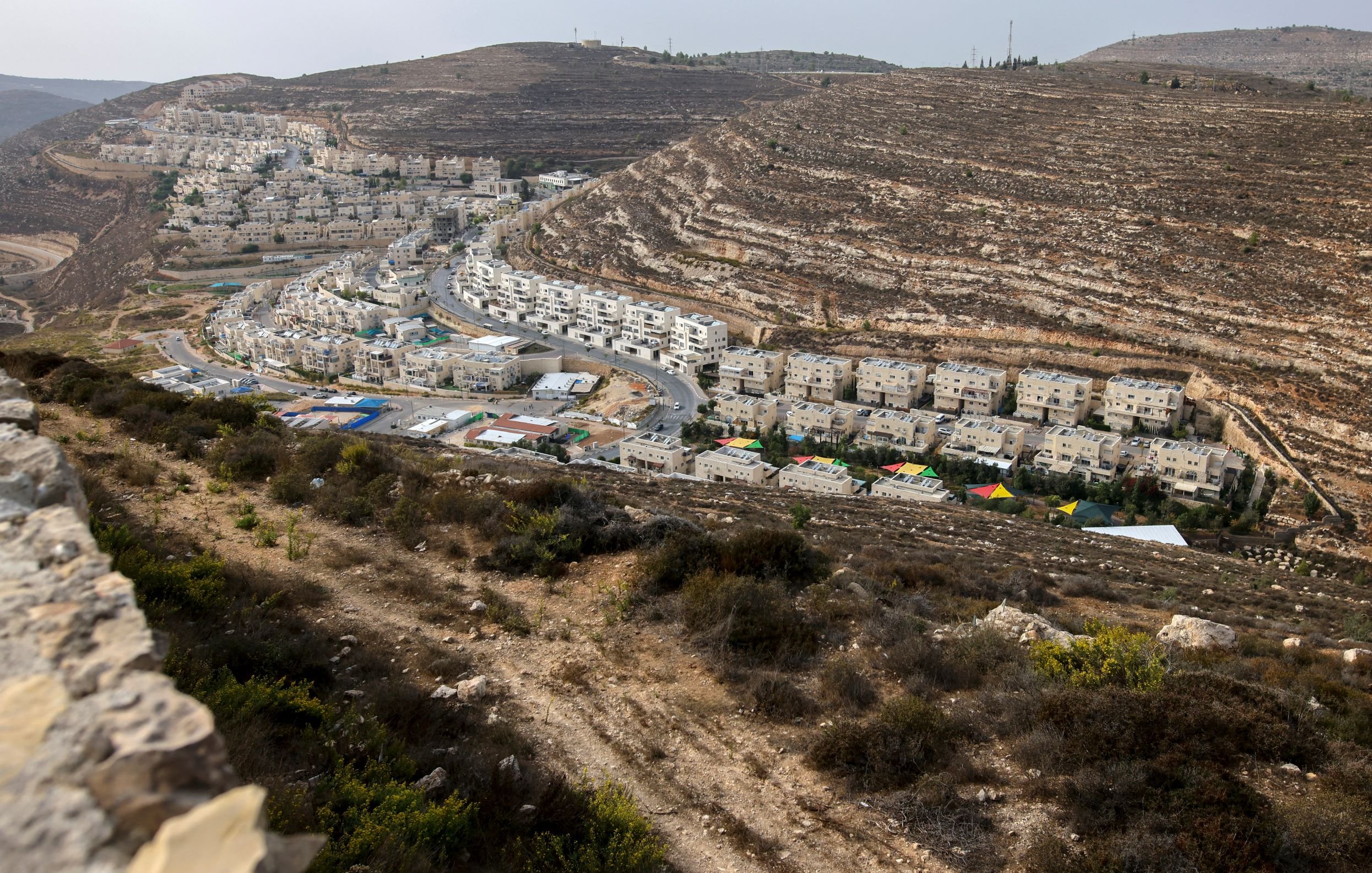 L'immagine mostra un paesaggio collinare, caratterizzato da una zona abitata con edifici bianchi disposti lungo una strada che si snoda tra le colline. In primo piano, si vede una vegetazione bassa e cespugliosa. Sullo sfondo, le colline presentano strati di roccia esposti, creando un effetto di stratificazione geologica. La luce appare naturale, suggerendo un'ora del giorno in cui il sole è alto, illuminando la scena con tonalità calde.