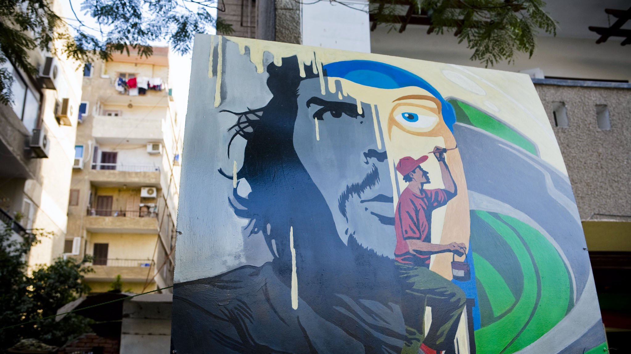 L'immagine mostra un murale artistico su un muro esterno. Il dipinto raffigura il volto di un uomo con un'espressione intensa, accanto a un artista in azione, intento a dipingere. Le tonalità utilizzate sono vivaci, con predominanza di blu e verde, creando un contrasto interessante. Sullo sfondo, si intravedono elementi architettonici e tende, suggerendo un ambiente urbano. Questo murale esprime una fusione tra arte e quotidianità.