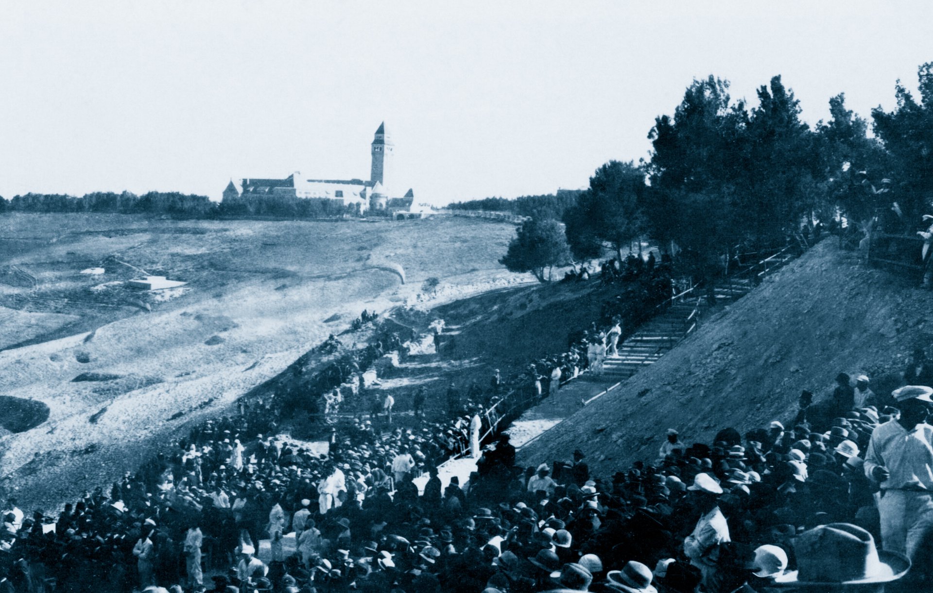 Un grande raduno di persone all'aperto, con un edifico distante e alberi circostanti.