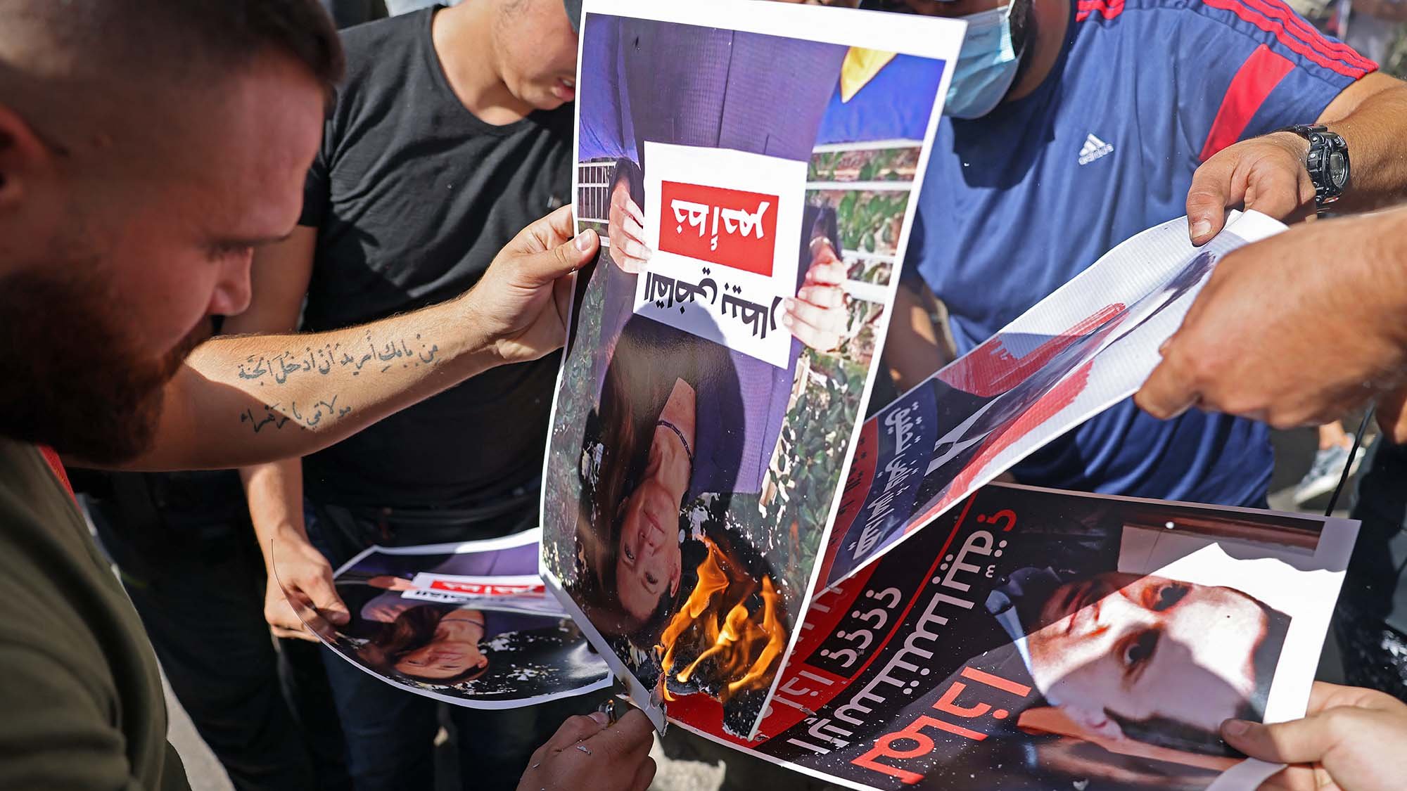 Nell'immagine si vedono diverse persone che stanno bruciando dei manifesti. I manifesti ritraggono volti, probabilmente di una figura pubblica, e sono esposti a una fiamma. Le persone sembrano partecipare attivamente a questa protesta o atto simbolico, esprimendo il loro discontento. L'atmosfera è di intensa emozione e opposizione.