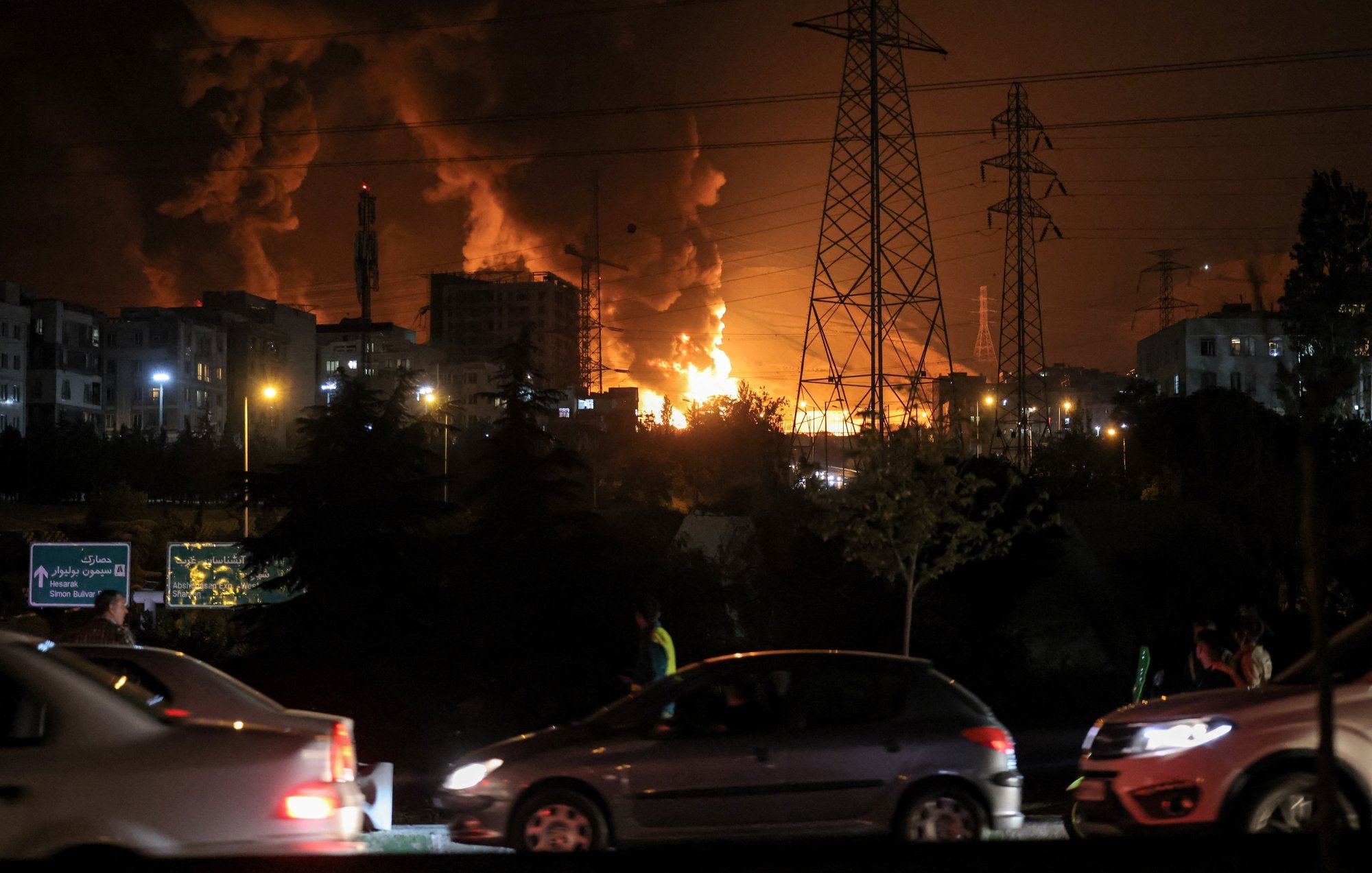 Un forte incendio illumina il cielo notturno, con fumi neri e auto in movimento.