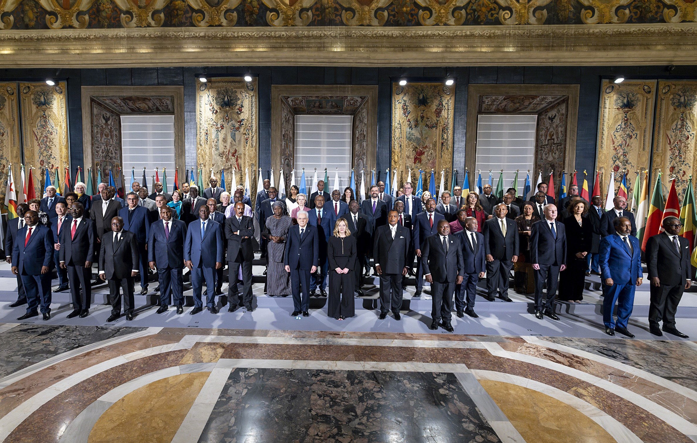 L'immagine mostra un gruppo di persone in un ambiente ufficiale, probabilmente durante un incontro o una conferenza internazionale. Si vedono diversi uomini e donne in abiti formali, che rappresentano vari paesi, dato che ci sono bandiere nazionali esposte sullo sfondo. L'atmosfera sembra seria e solenne, con tutti i partecipanti schierati in modo ordinato. Le decorazioni e l'architettura dell'ambiente circostante danno un tocco di grandezza all'evento.