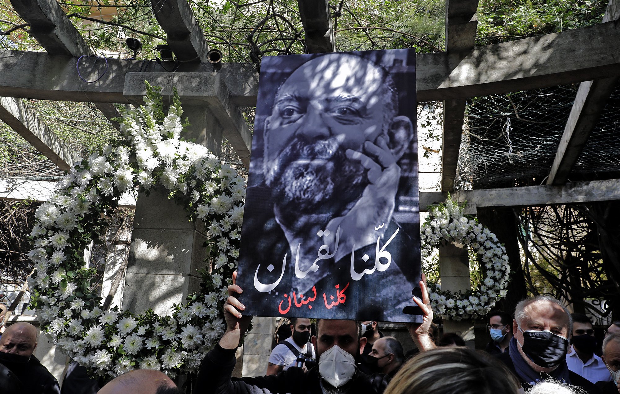 L'immagine mostra una persona che tiene in alto un grande cartello. Il cartello presenta un ritratto in bianco e nero di un uomo con una barba, accompagnato da un testo in arabo che significa "Siamo tutti Libano". Sullo sfondo, ci sono decorazioni floreali e un ambiente che sembra essere un luogo commemorativo. La scena trasmette un senso di unità e solidarietà.