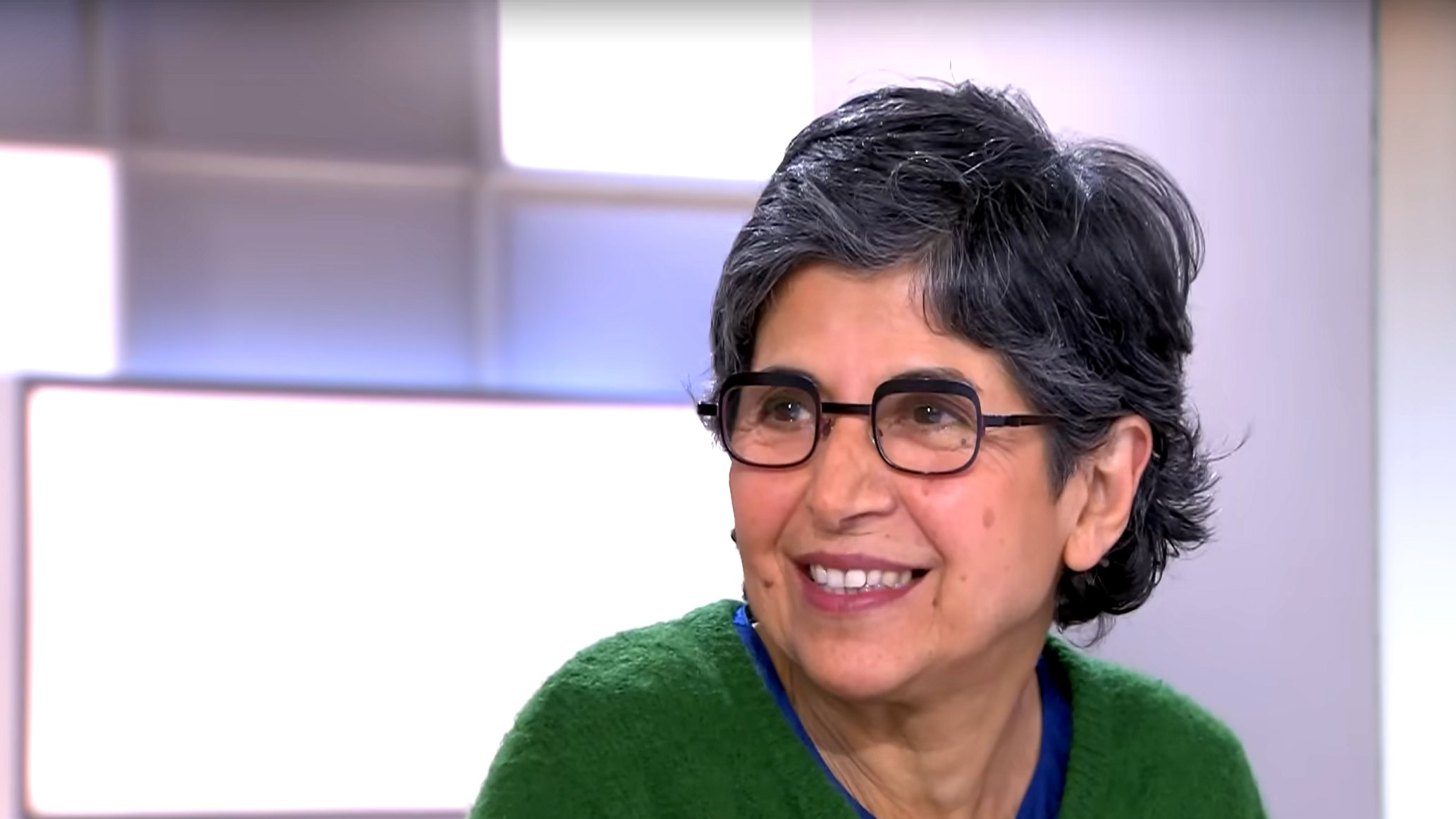 L'immagine mostra una donna con capelli grigi e ondulati, indossando occhiali e un maglione verde. Sembra sorridere e avere un'espressione accogliente. Sullo sfondo ci sono sfumature chiare che danno un'atmosfera luminosa e positiva.