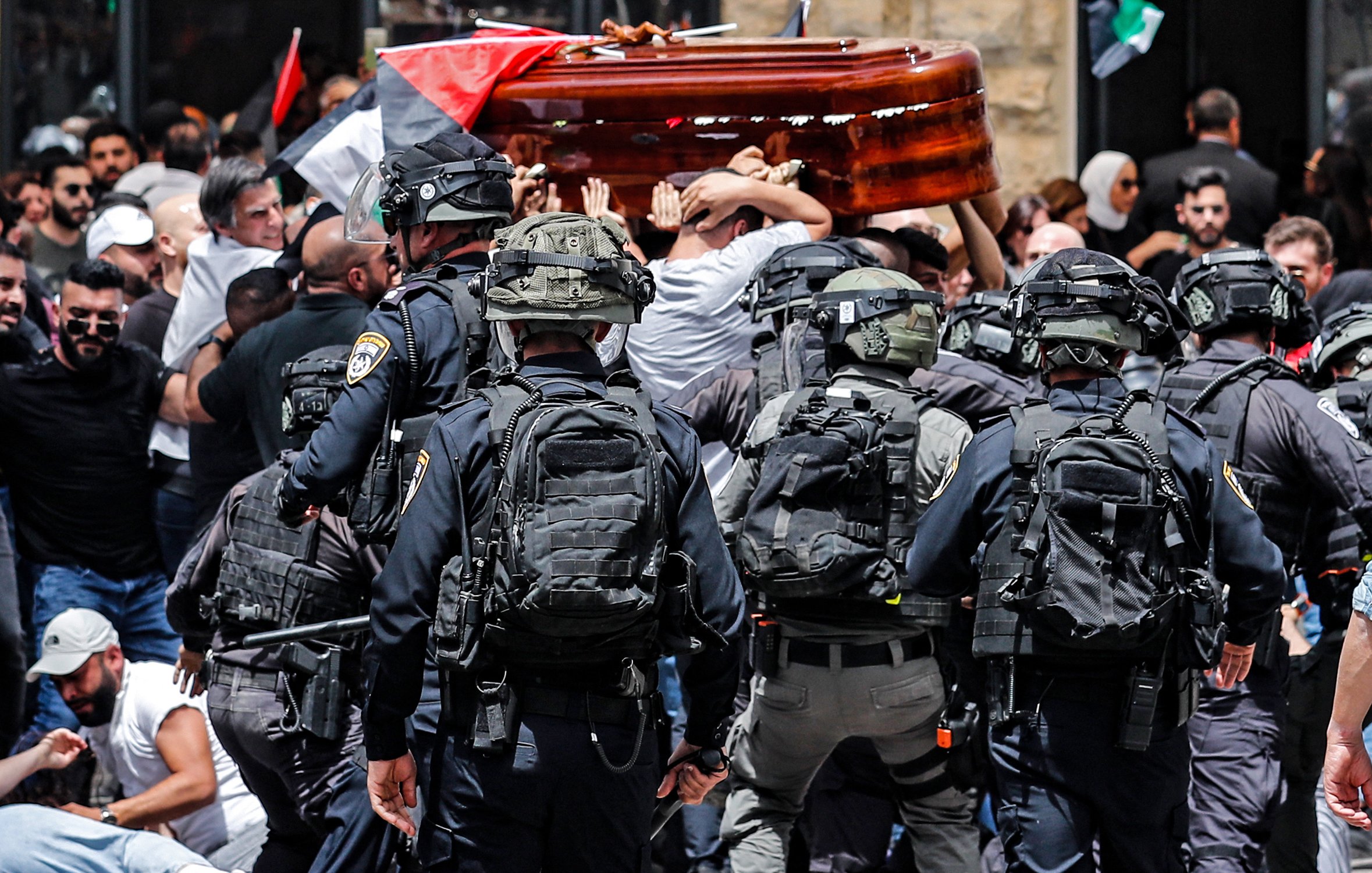 L'immagine rappresenta una scena di tensione durante una manifestazione. Ci sono persone che trasportano un barella, probabilmente durante un funerale o una commemorazione, mentre un gruppo di agenti di polizia in equipaggiamento anti-sommossa sta cercando di mantenere l'ordine. Molti manifestanti sono visibili, con bandiere che simboleggiano le loro rivendicazioni, e l'atmosfera appare carica di emozione e conflitto. La scena è caratterizzata da un forte senso di mobilitazione e protesta.