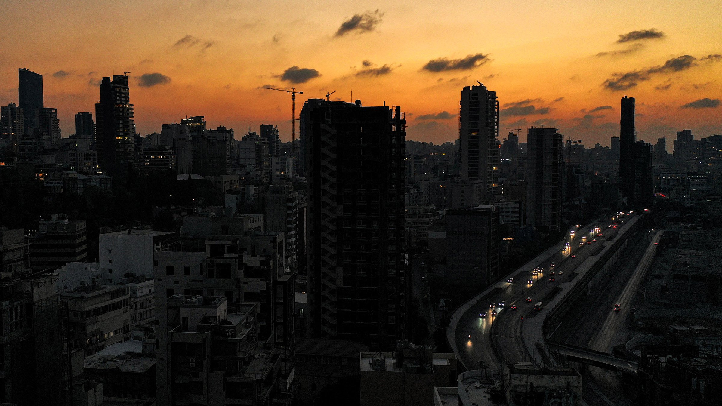 L'immagine ritrae un paesaggio urbano al tramonto, con il cielo che mostra tonalità calde di arancione e giallo. Silhouette di grattacieli si stagliano contro il cielo, creando un contrasto tra il buio degli edifici e la luce del tramonto. In basso, si vede una strada con auto che circolano, illuminata dalle luci dei veicoli. L'atmosfera è serena e suggestiva, tipica di un momento di transizione tra giorno e notte.