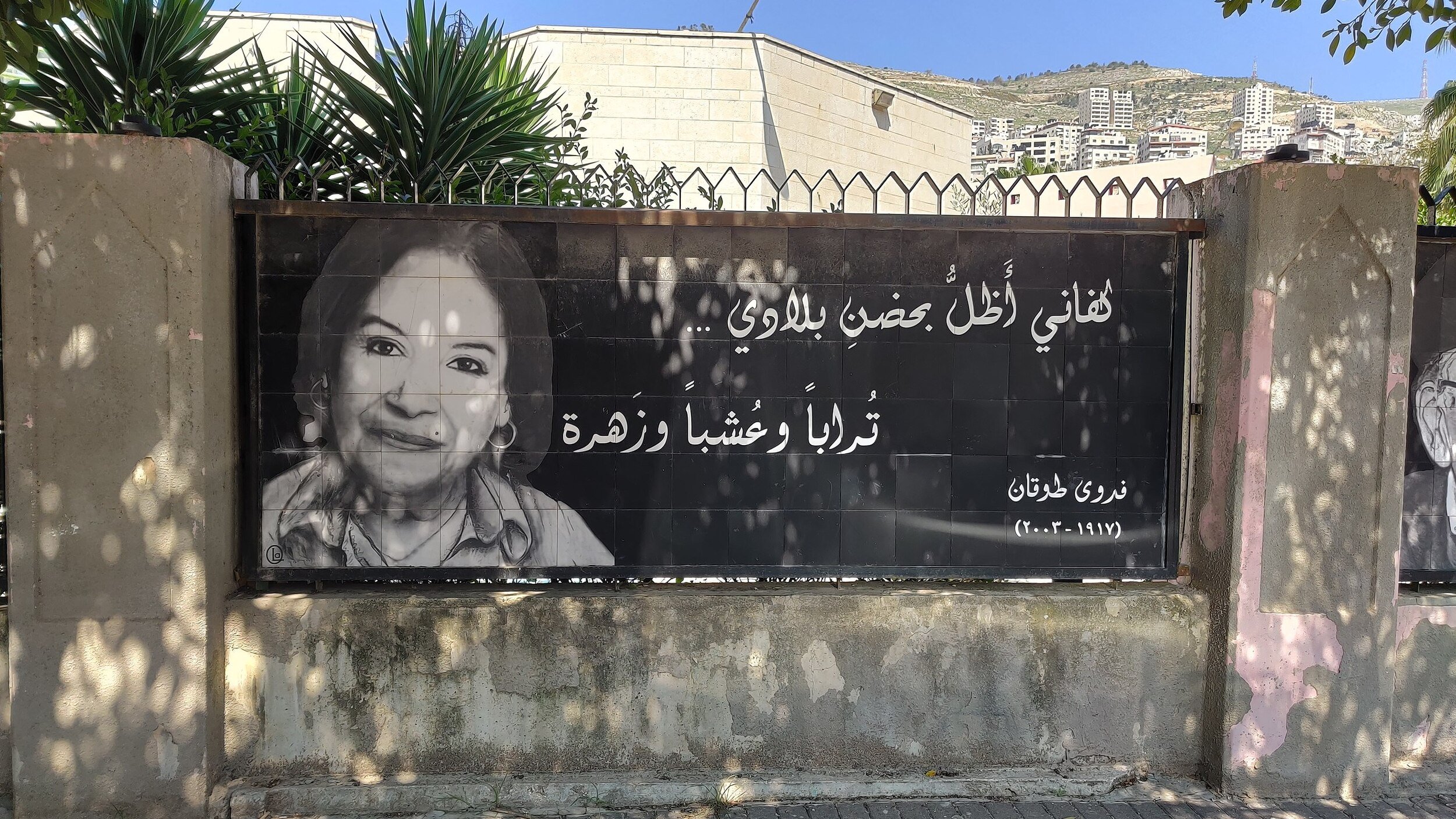 L'immagine mostra un grande striscione che riporta una fotografia di una donna sorridente, accompagnata da un testo in arabo. Il contesto sembra essere un'area urbana, con edifici sullo sfondo e piante verdi nelle vicinanze. L'atmosfera è accogliente e il messaggio sullo striscione appare positivo e invitante.