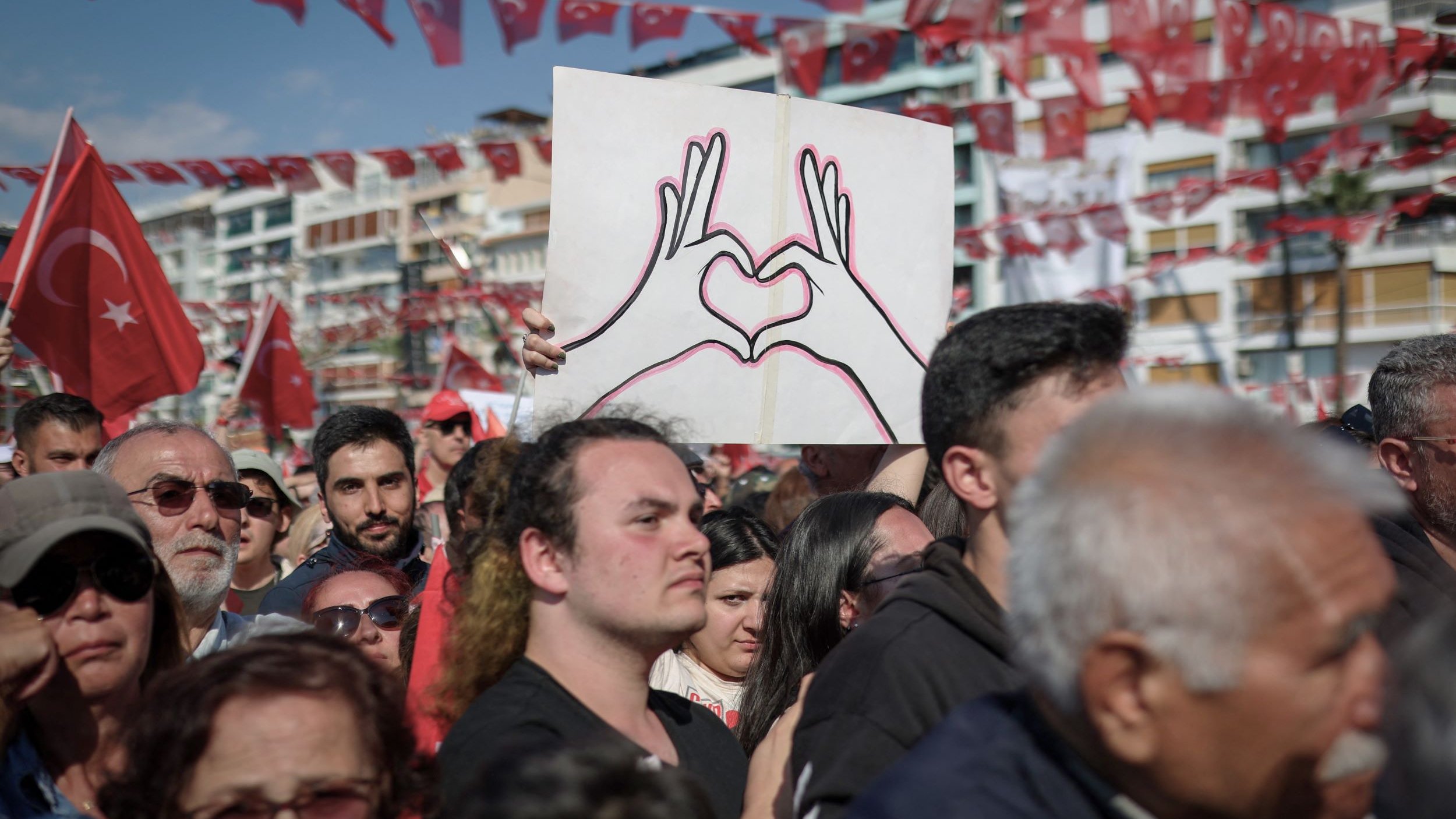 L'immagine mostra una grande folla riunita in un evento all'aperto, probabilmente una manifestazione o una celebrazione. Alcuni partecipanti sostengono bandiere turche, che sono di un rosso acceso, mentre un uomo in primo piano è visibile con una maglietta nera e guarda verso la telecamera. Dietro di lui, c'è un cartello che rappresenta due mani che formano un cuore, simbolo di amore o unità. L'atmosfera sembra essere festosa e piena di energia, con molte persone che condividono un momento di comunità.