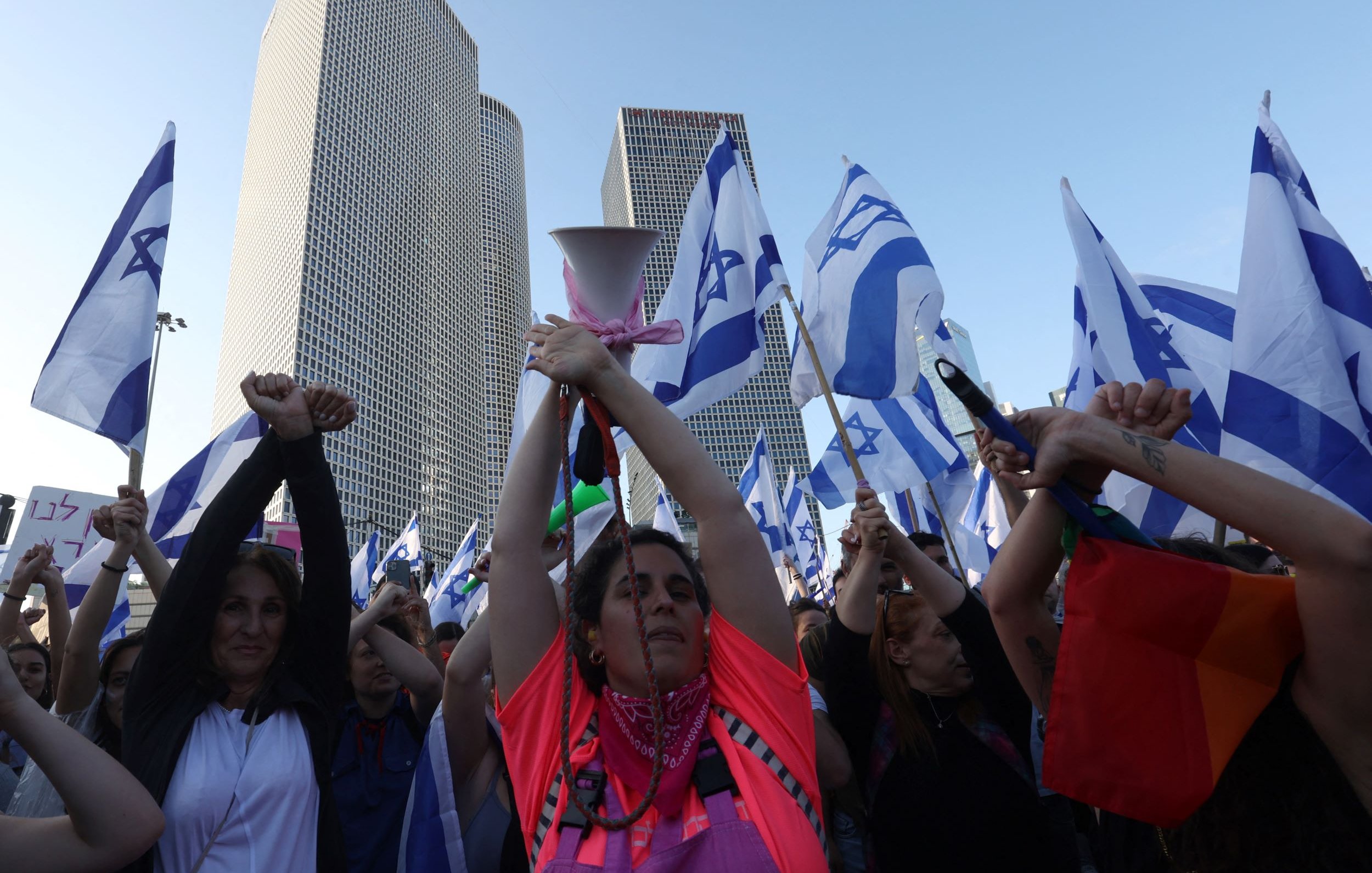 L'immagine mostra una folla di persone che partecipa a una manifestazione. Alcuni partecipanti tengono in alto delle bandiere israeliane, mentre molti altri alzano le mani, dimostrando un senso di unità e protesta. In primo piano, si può vedere una donna con una maglietta di colore neon che sembra guidare la manifestazione con un megafono. Sullo sfondo, si notano alti edifici moderni, suggerendo che l'evento si svolge in un contesto urbano. L'atmosfera è carica di energia e determinazione.