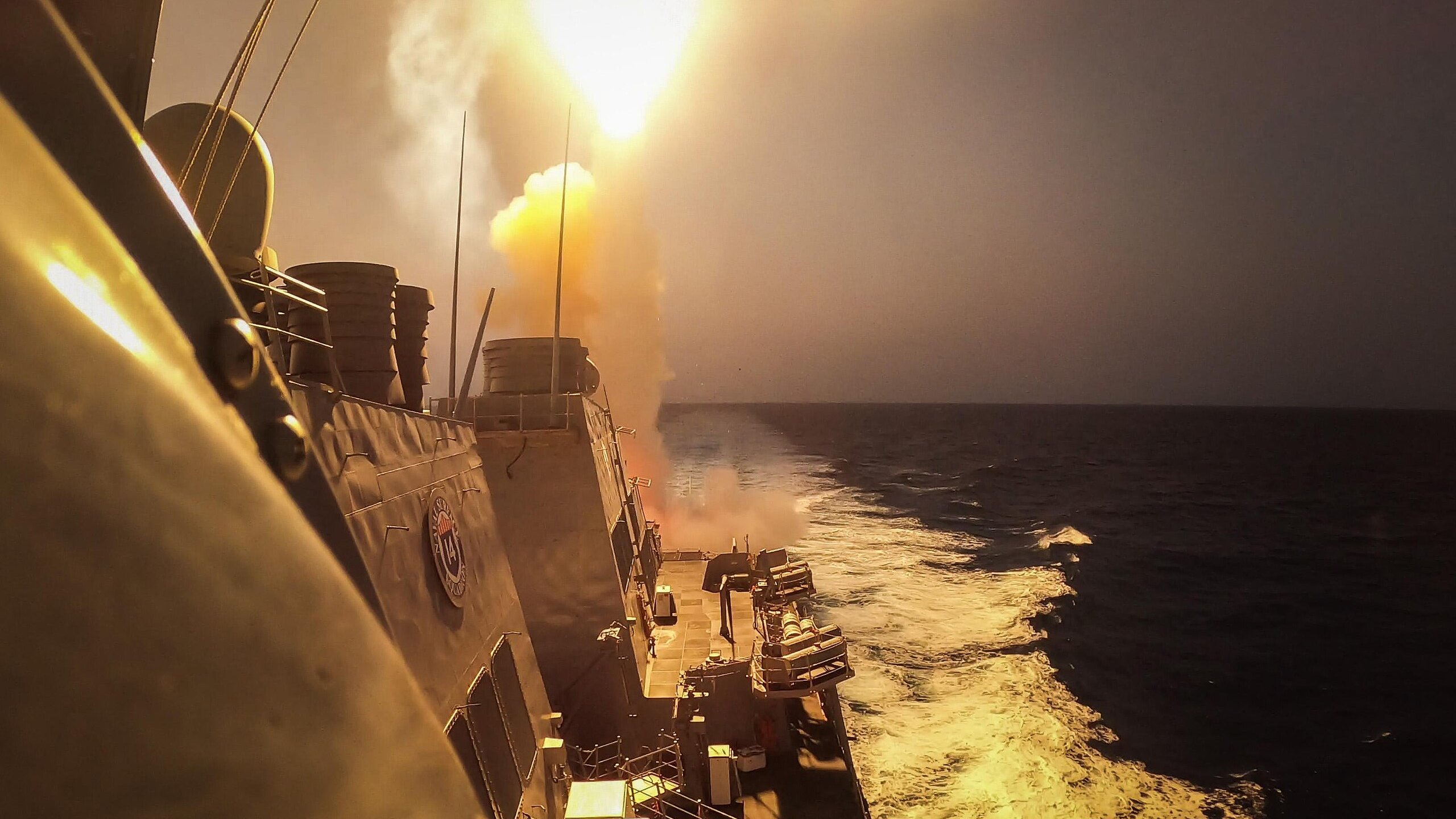 L'immagine mostra una scena marittima in cui un missile viene lanciato da una nave da guerra. Si vede il missile che viene sparato con un forte bagliore e un'esplosione di fumi dietro di esso. In primo piano si possono notare le strutture della nave e il mare in movimento, evidenziato da schiuma bianca. Il cielo appare velato e si intuisce un momento di grande intensità e potenza militare.