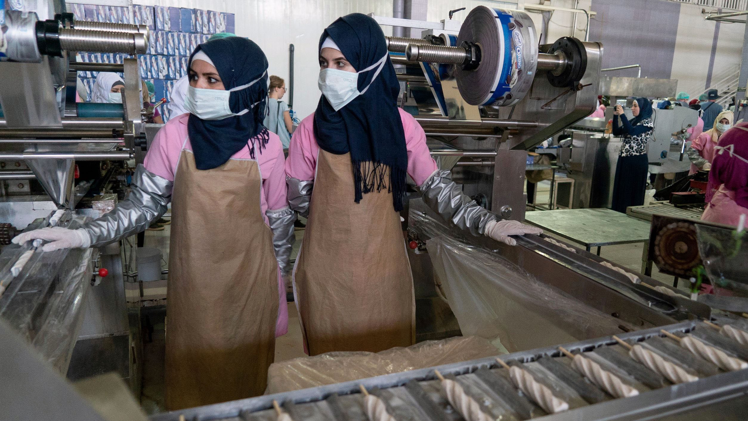 L'immagine mostra due donne in un ambiente di lavoro, probabilmente in una fabbrica. Indossano abiti protettivi, tra cui mascherine e foulard, e stanno operando a vicino a una macchina industriale. Sullo sfondo si possono intravedere altre persone e diversi macchinari, suggerendo un'ambientazione dedicata alla produzione. L'atmosfera appare professionalmente orientata e organizzata.