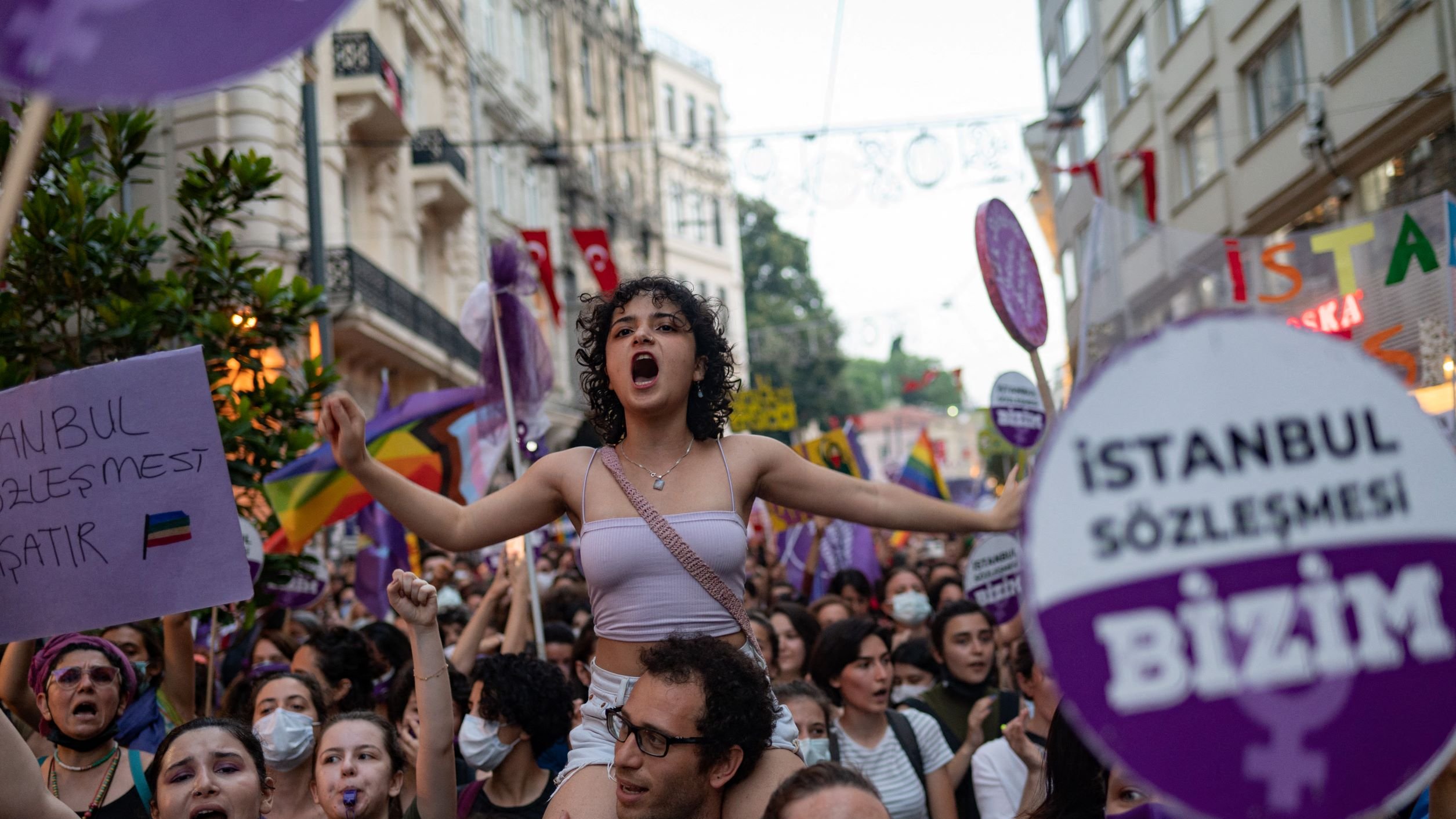 L'immagine mostra un gruppo di persone in una manifestazione per i diritti, con un forte focus sulle tematiche femministe e LGBTQ+. Una giovane donna, in primo piano e con i capelli ricci, è in piedi sulla spalle di un'altra persona e grida, esprimendo la sua passione. Molti partecipanti, prevalentemente donne, sono circondati da bandiere e cartelli colorati, alcuni con slogan che richiamano l'attenzione su temi di uguaglianza e diritti umani. L'atmosfera è vibrante e carica di energia, con un chiaro senso di solidarietà tra i manifestanti.